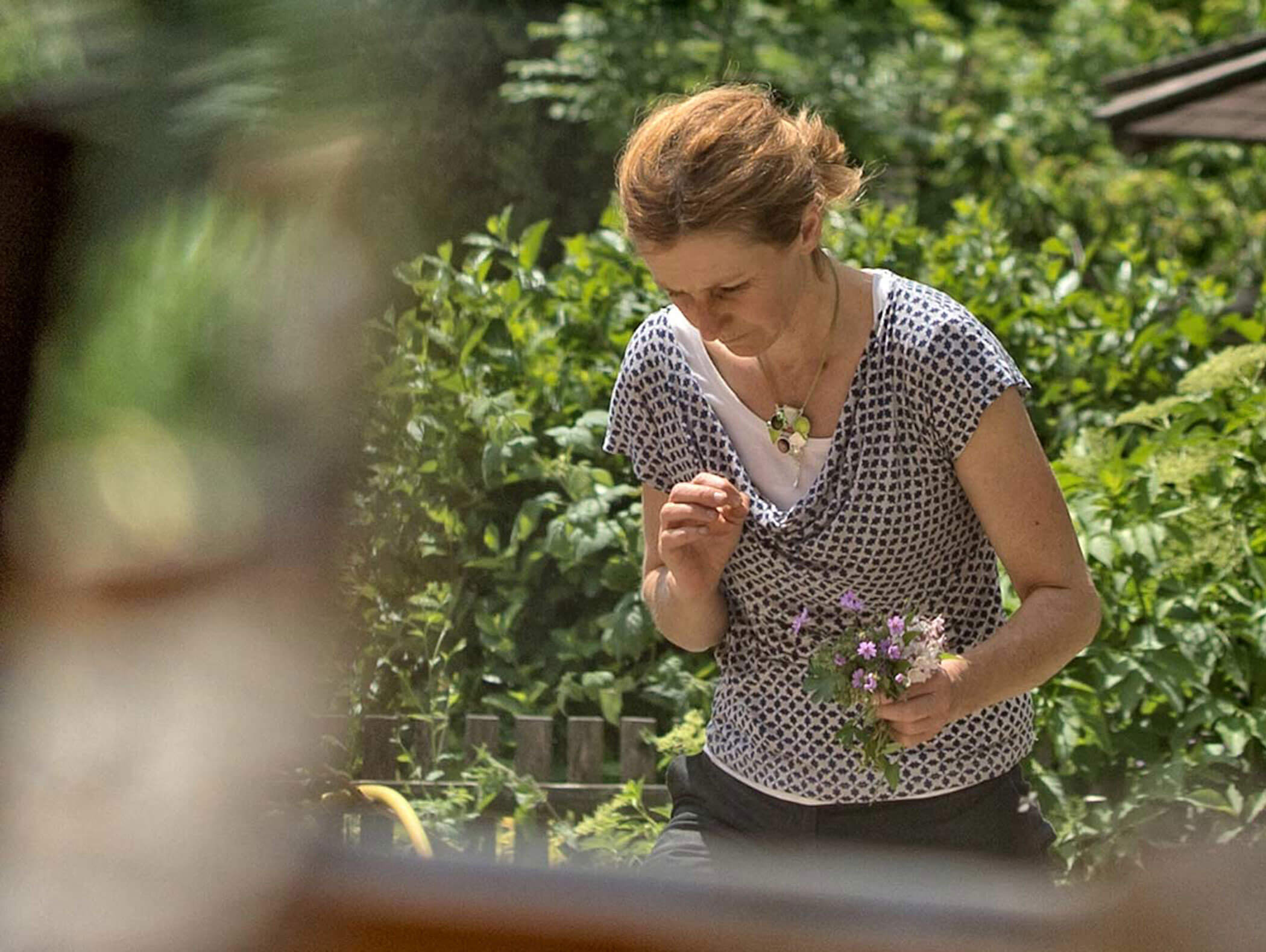 Donna con capelli corti che tiene in mano piccoli fiori viola, in piedi all'aperto in un giardino con fogliame verde. - Hotel Moosmair