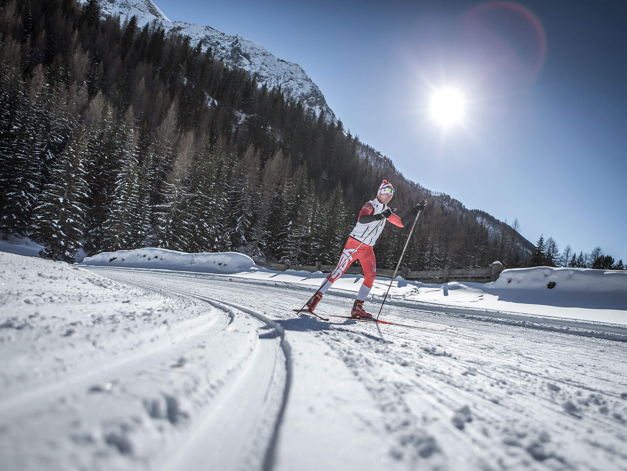 Uno sciatore di fondo scivola su piste innevate sotto un sole splendente con montagne e pini sullo sfondo. - Hotel Moosmair
