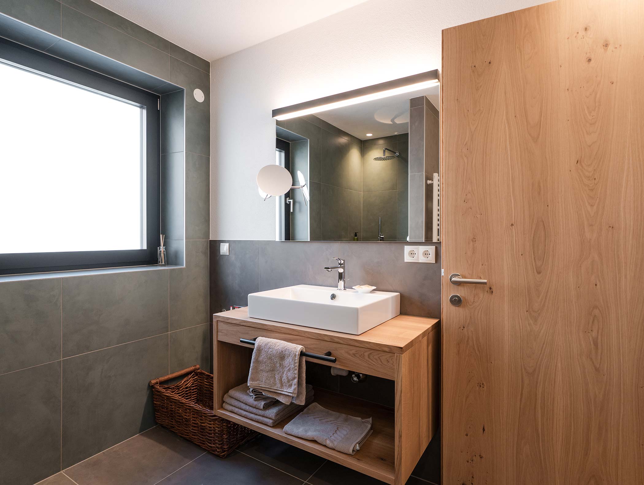 Bagno moderno con piastrelle grigie, lavabo in legno, lavabo a vaschetta, grande specchio e un cesto di vimini con gli asciugamani. - Hotel Moosmair