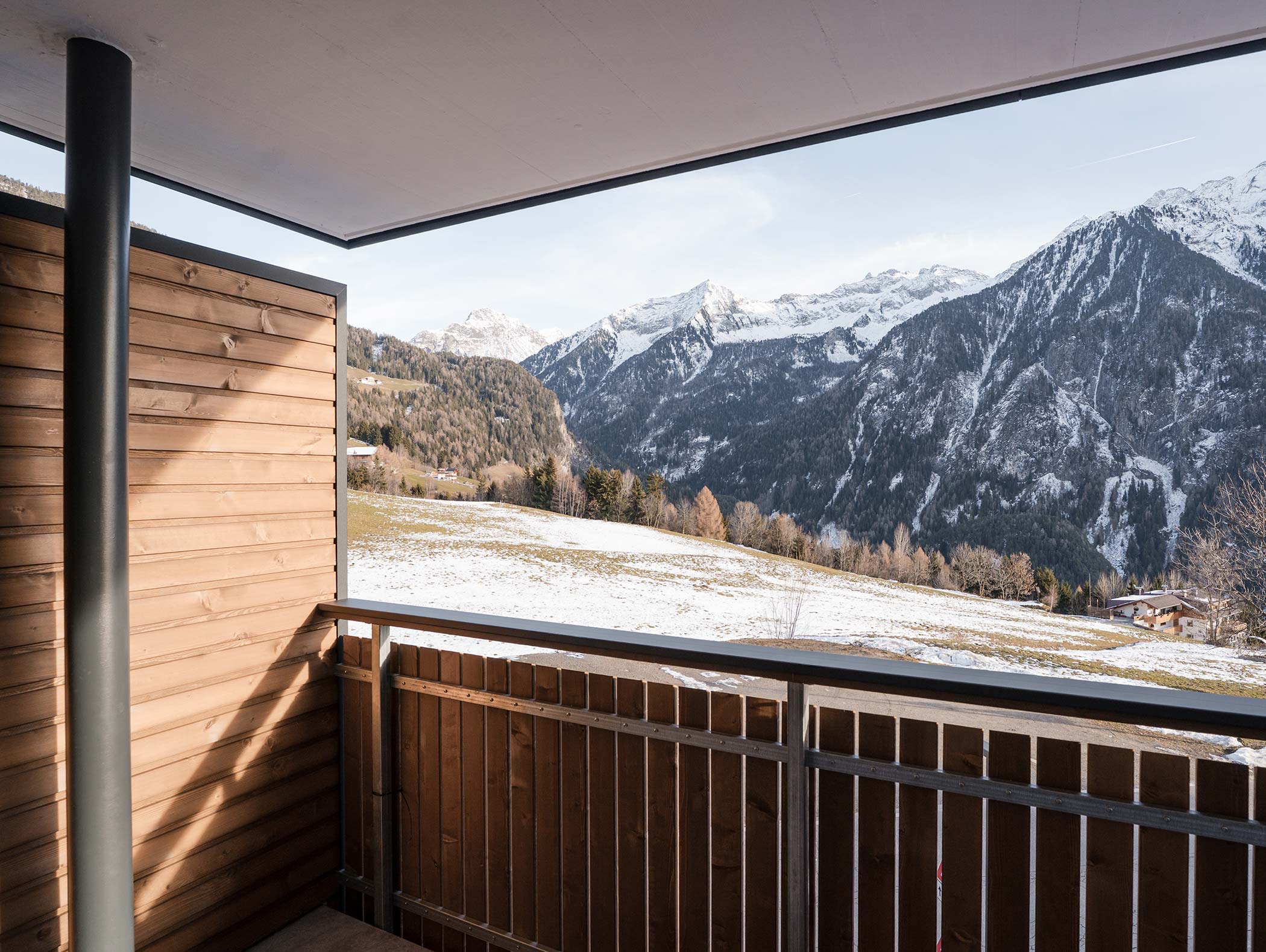 Un balcone in legno si affaccia sulle montagne innevate e su un campo erboso sotto un cielo parzialmente nuvoloso. - Hotel Moosmair