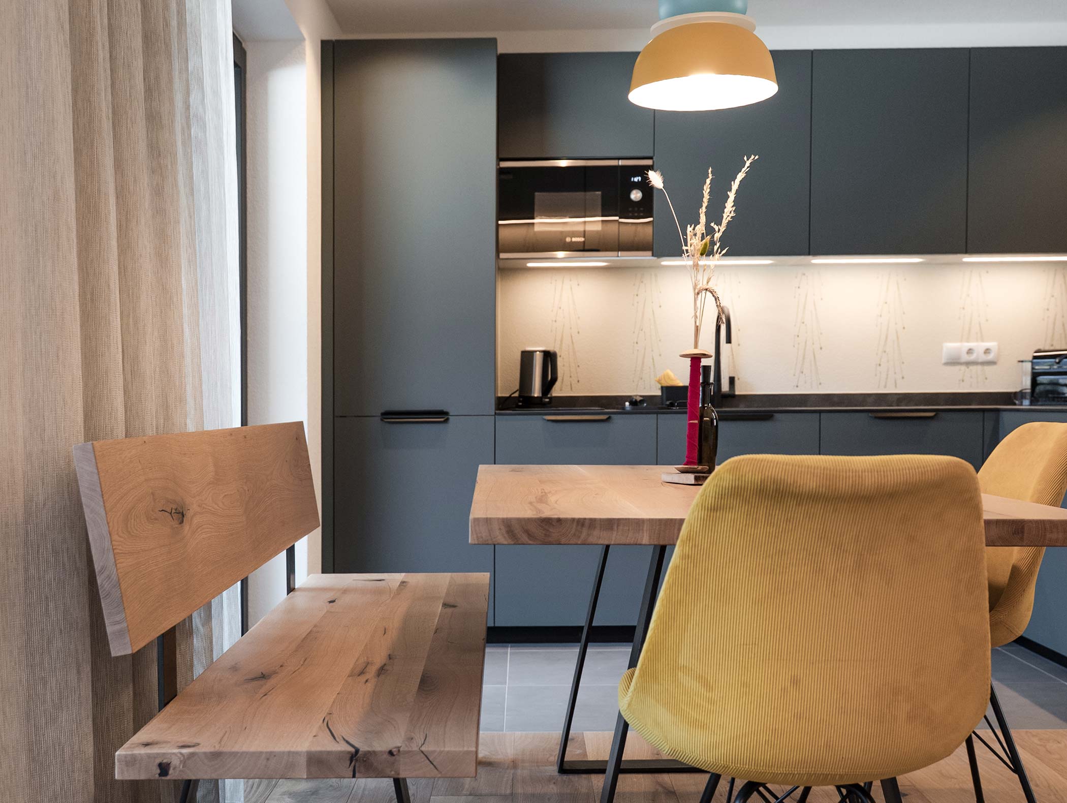 Cucina moderna con armadietti opachi, tavolo in legno, sedie gialle e panca con illuminazione a sospensione. - Hotel Moosmair