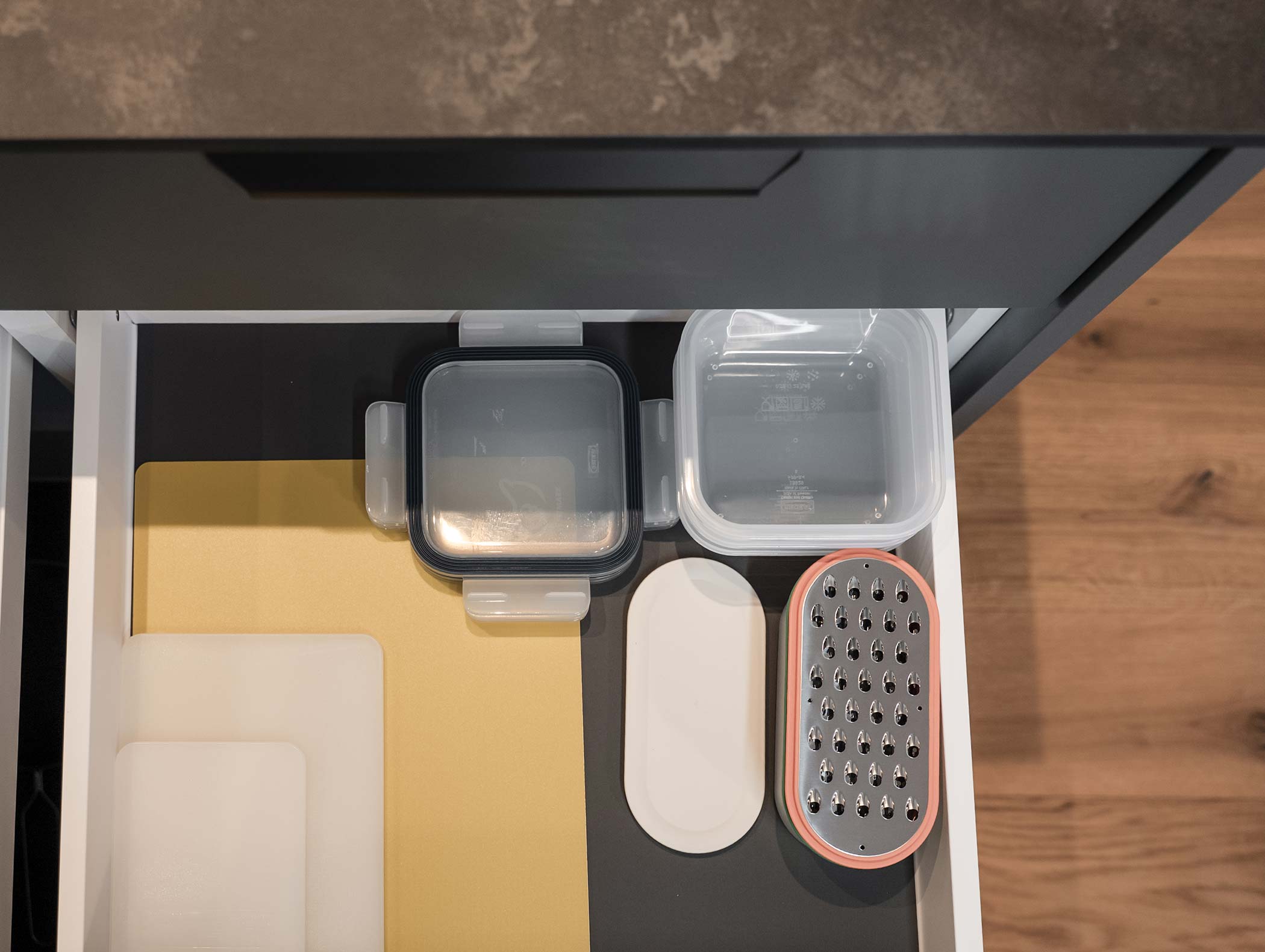 Cassetto da cucina aperto con taglieri, contenitori per alimenti e una grattugia di metallo ordinatamente organizzati all'interno. - Hotel Moosmair