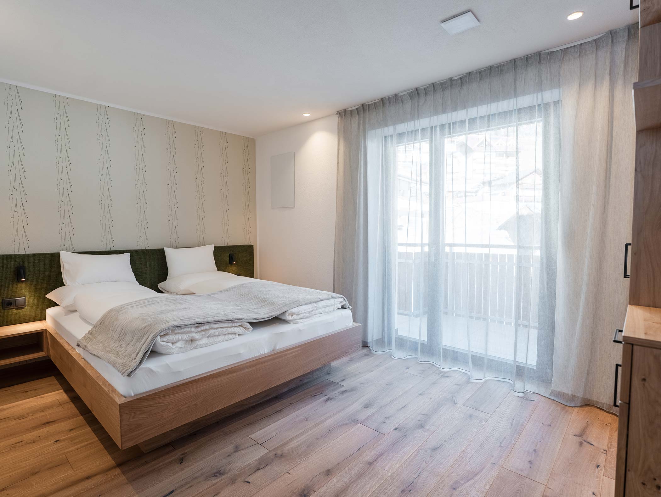 Camera da letto moderna con letto matrimoniale, biancheria da letto chiara, pavimento in legno e tende velate che coprono un'ampia finestra. - Hotel Moosmair