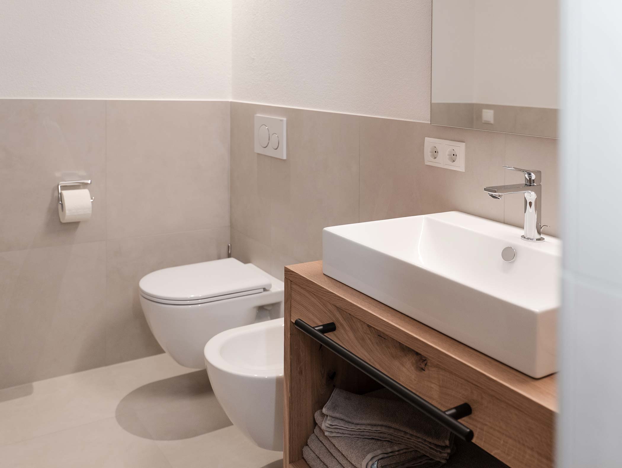 Bagno moderno con WC a parete, bidet, lavabo, mobiletto in legno, ripiano per asciugamani e porta carta igienica. - Hotel Moosmair