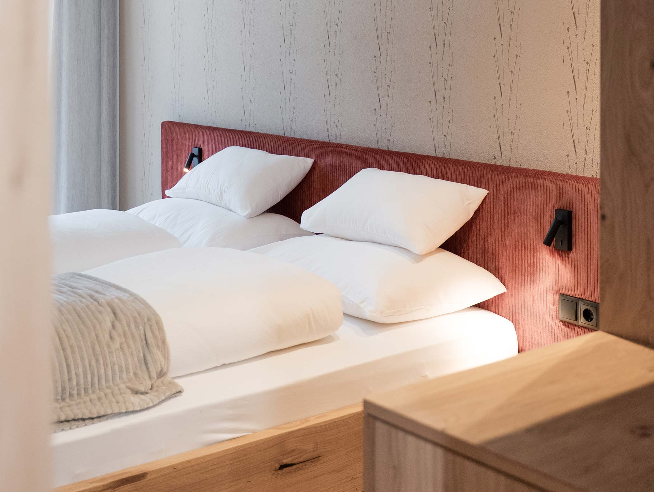 Un letto ben fatto con lenzuola bianche, testata rossa e luci da lettura a parete in una stanza luminosa. - Hotel Moosmair