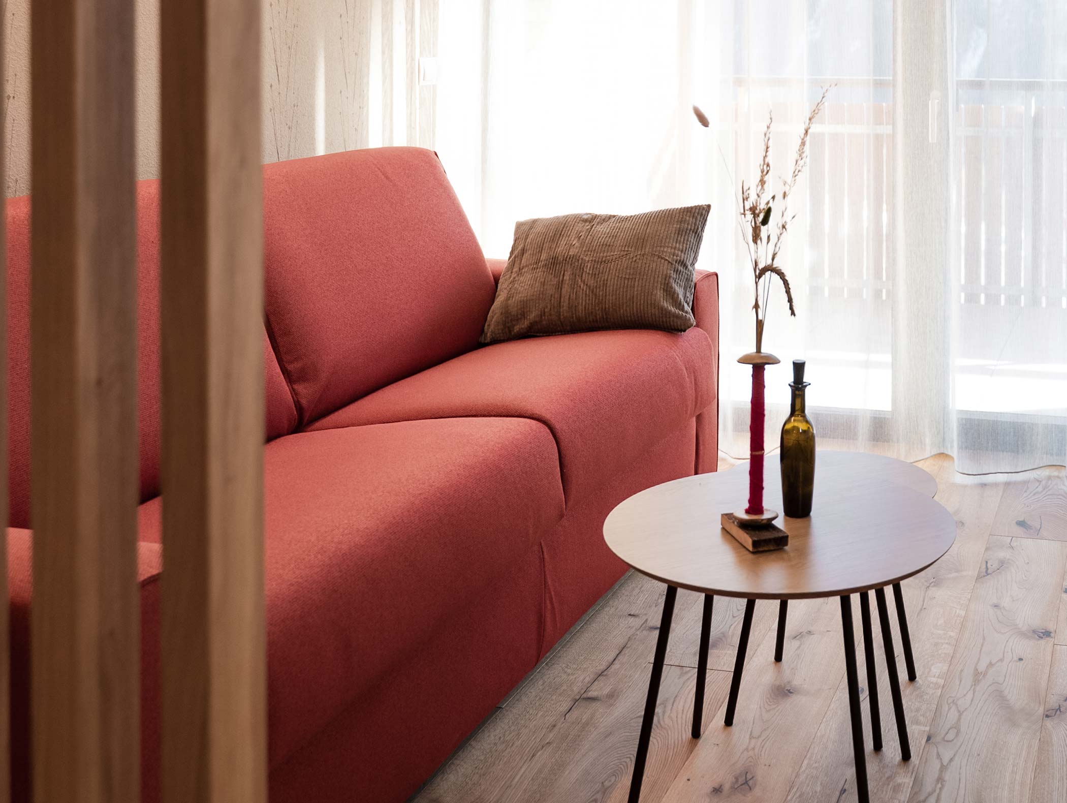 Divano rosso con cuscino marrone accanto a un tavolo rotondo di legno con vasi e candelieri, vicino a finestre illuminate dal sole. - Hotel Moosmair