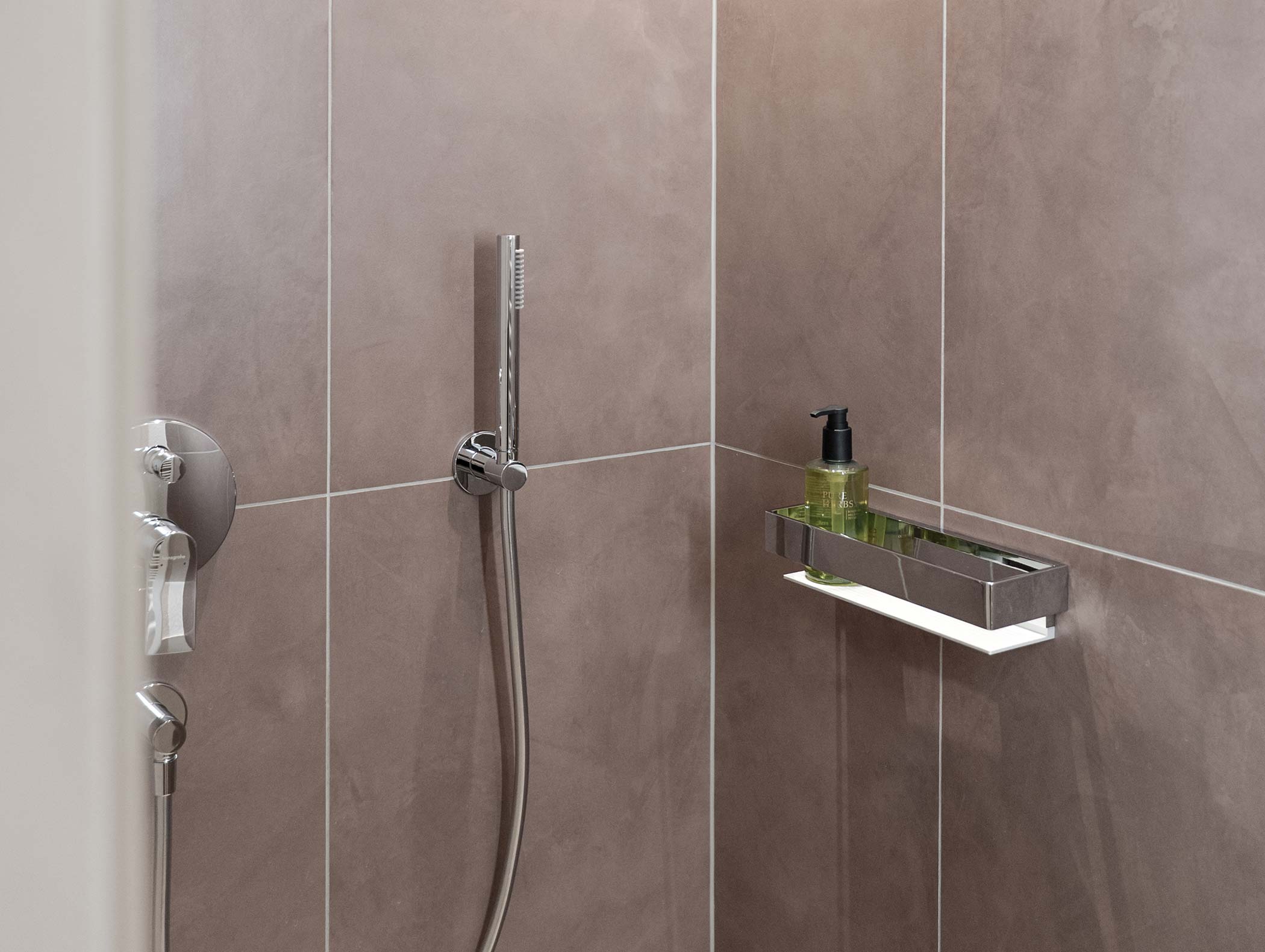 Doccia moderna con soffione a mano, mensola a muro e dispenser di sapone verde su pareti piastrellate marroni. - Hotel Moosmair