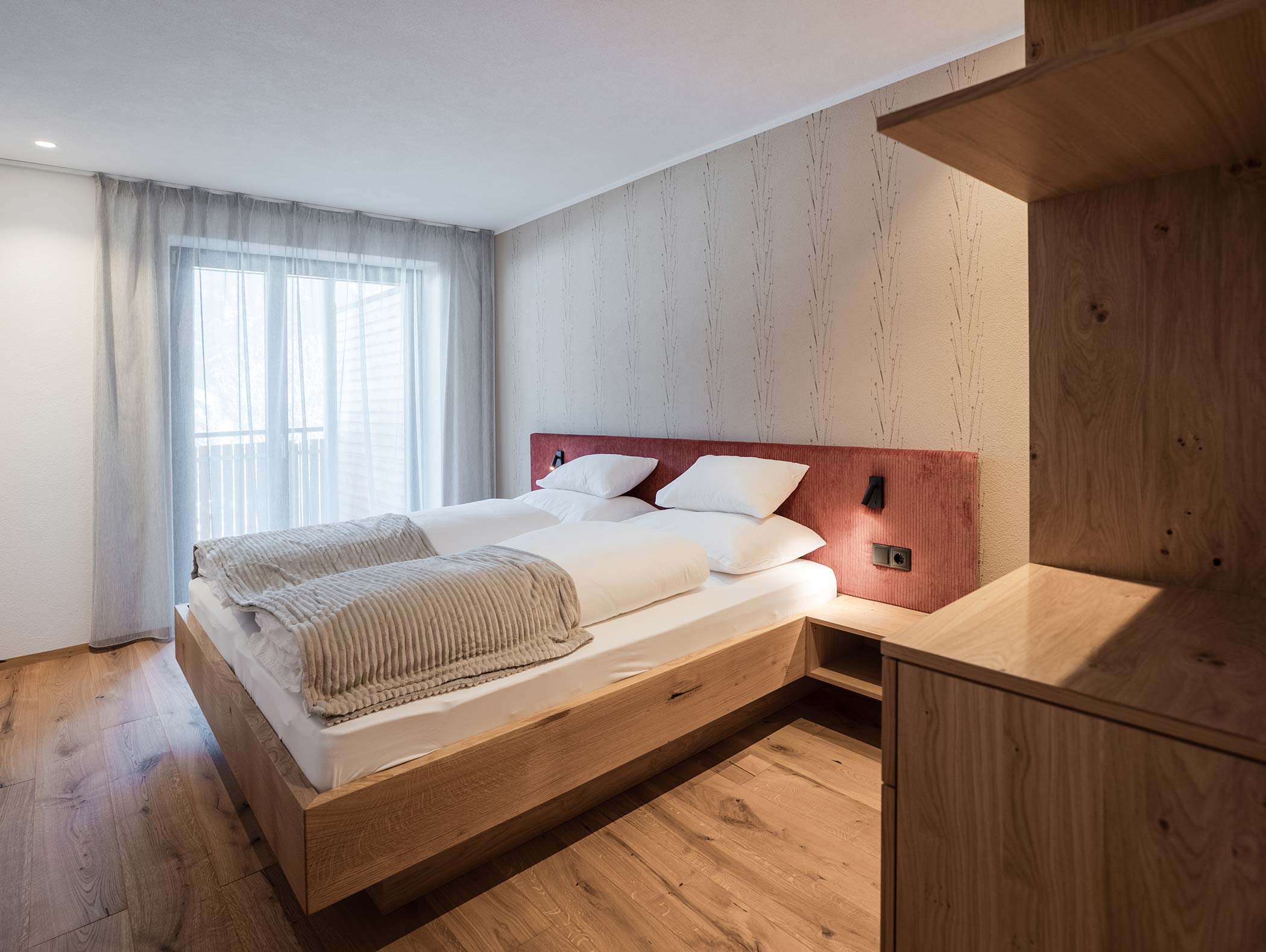 Camera moderna con letto matrimoniale, mobili in legno, ampia finestra con tende velate e luci soffuse. - Hotel Moosmair