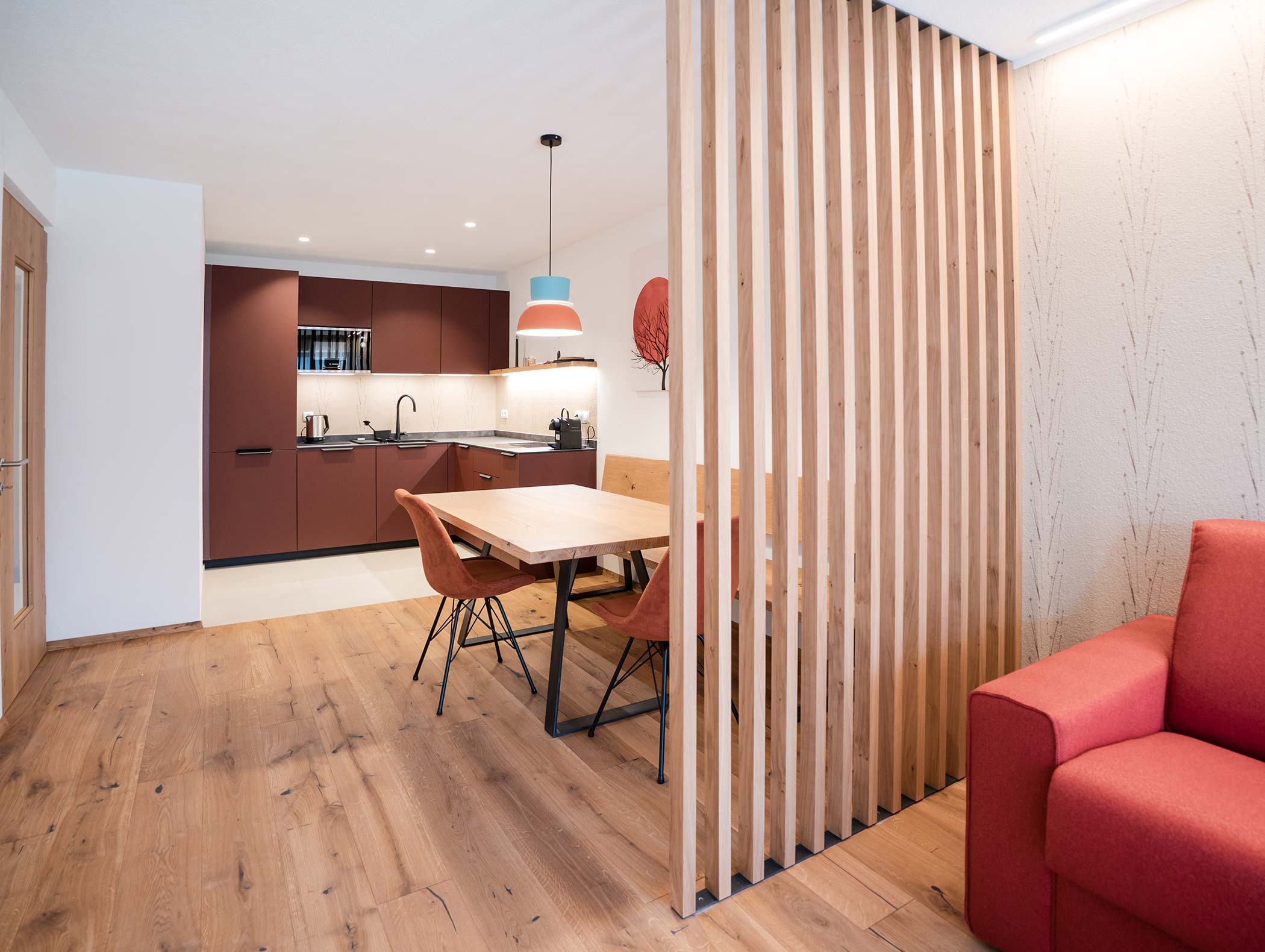 Cucina moderna e zona pranzo con pavimento in legno, divisorio a doghe e sedie rosse; divano rosso in primo piano. - Hotel Moosmair