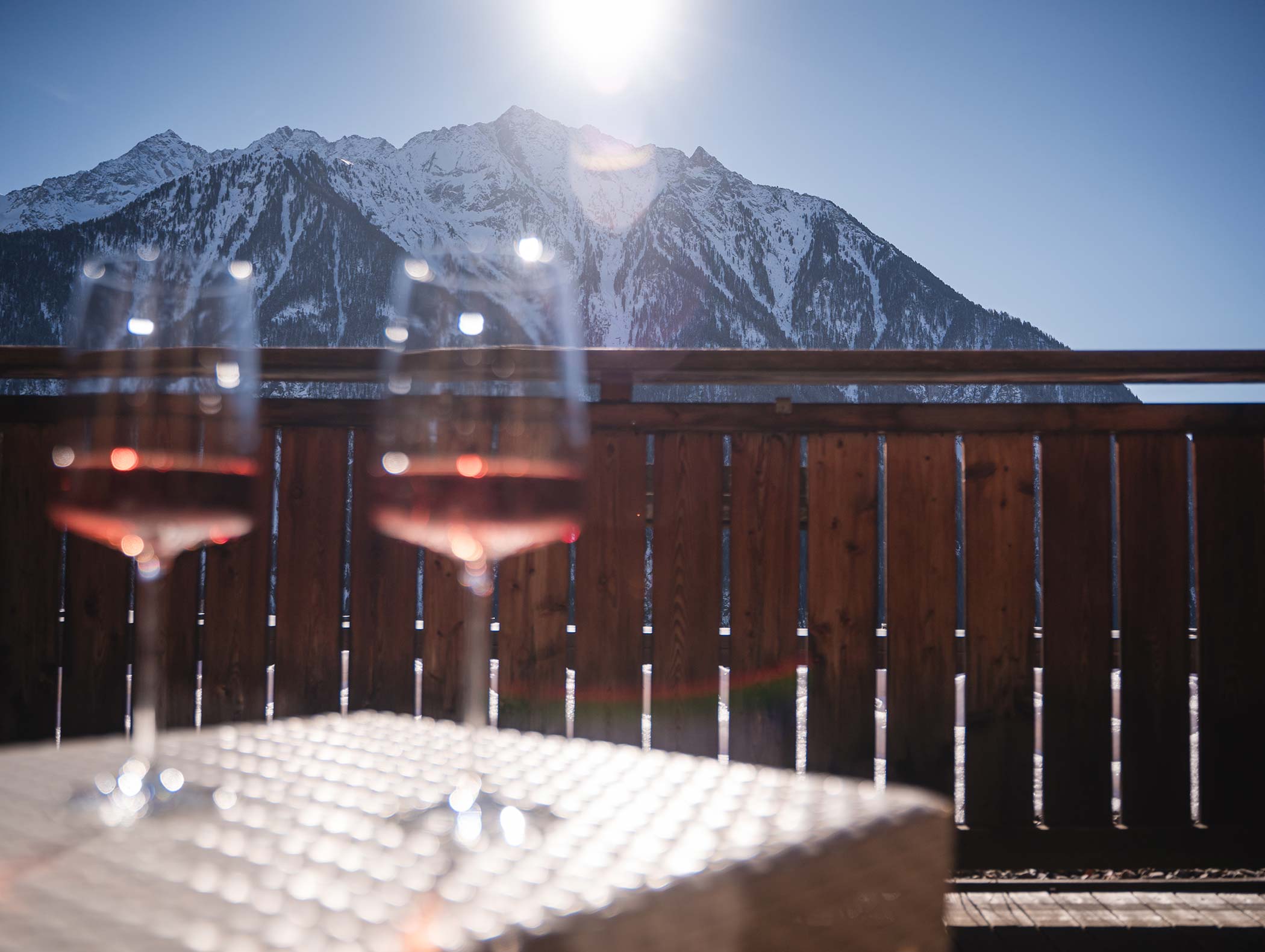 Due bicchieri di vino rosé su un tavolo con montagne innevate e un sole splendente sullo sfondo. - Hotel Moosmair