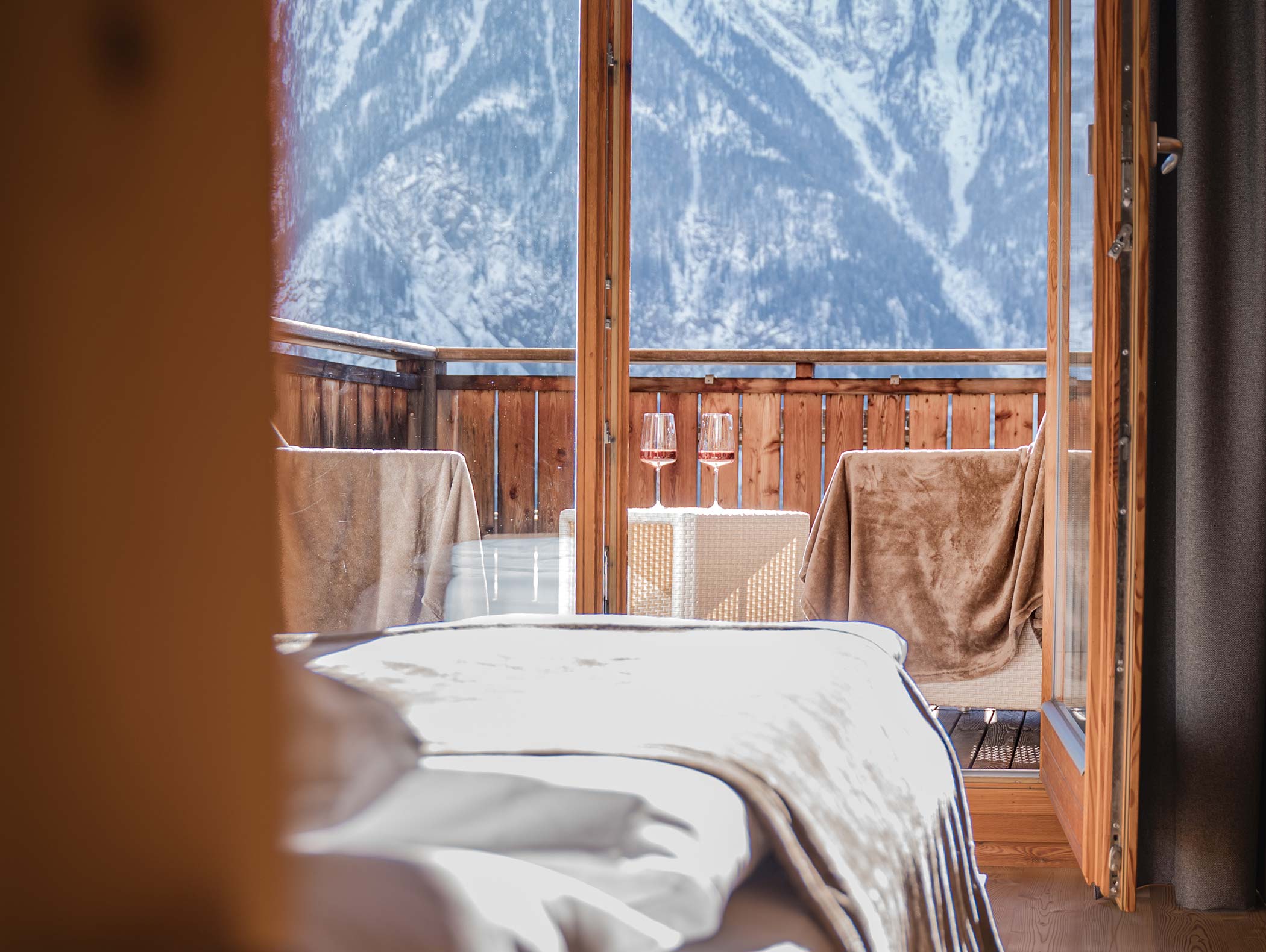 Camera accogliente con porte aperte su un balcone, due bicchieri di vino e montagne innevate sullo sfondo. - Hotel Moosmair