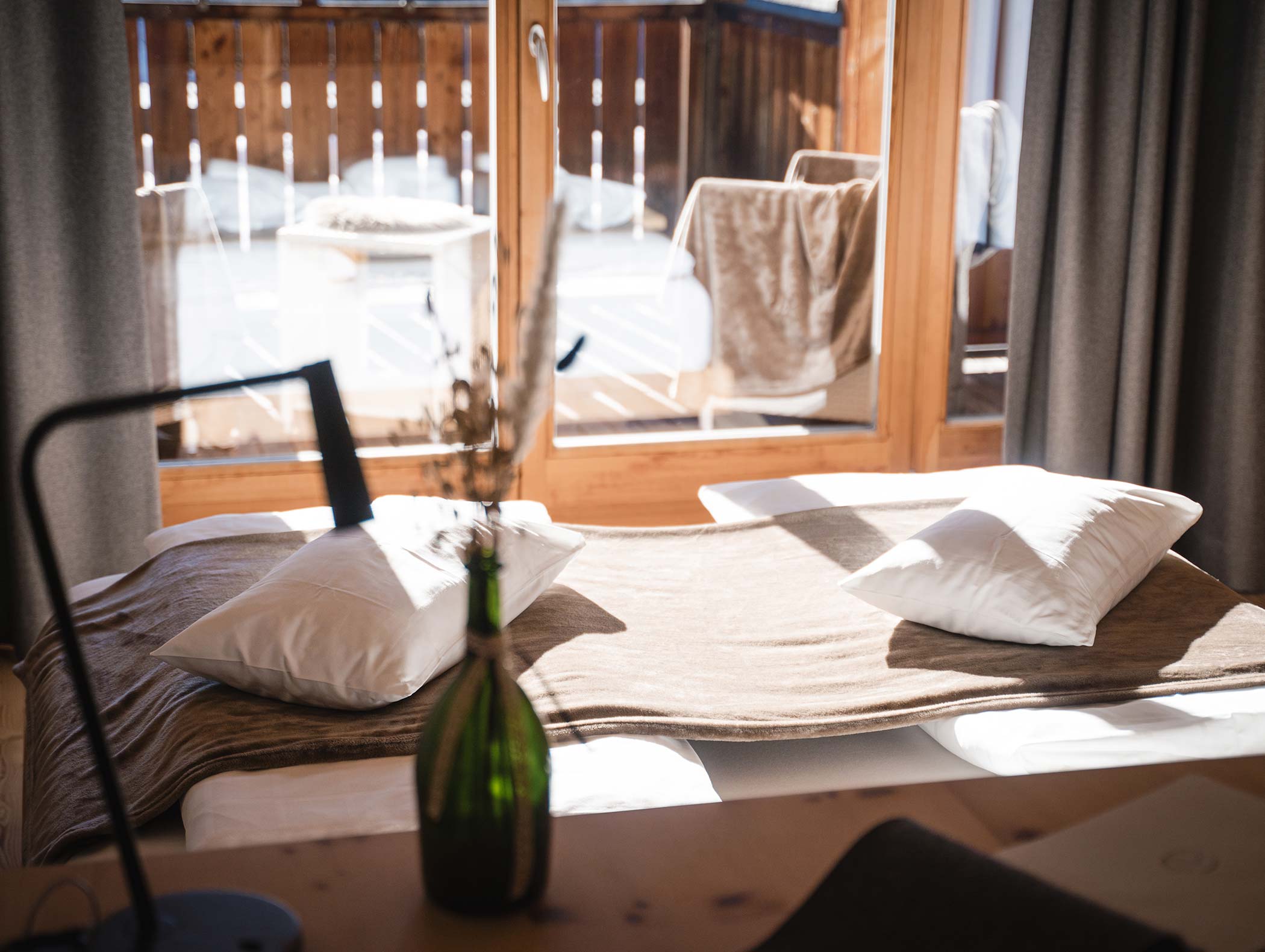 Accogliente camera da letto illuminata dal sole con due cuscini su un letto vicino a grandi finestre e un vaso verde in primo piano. - Hotel Moosmair