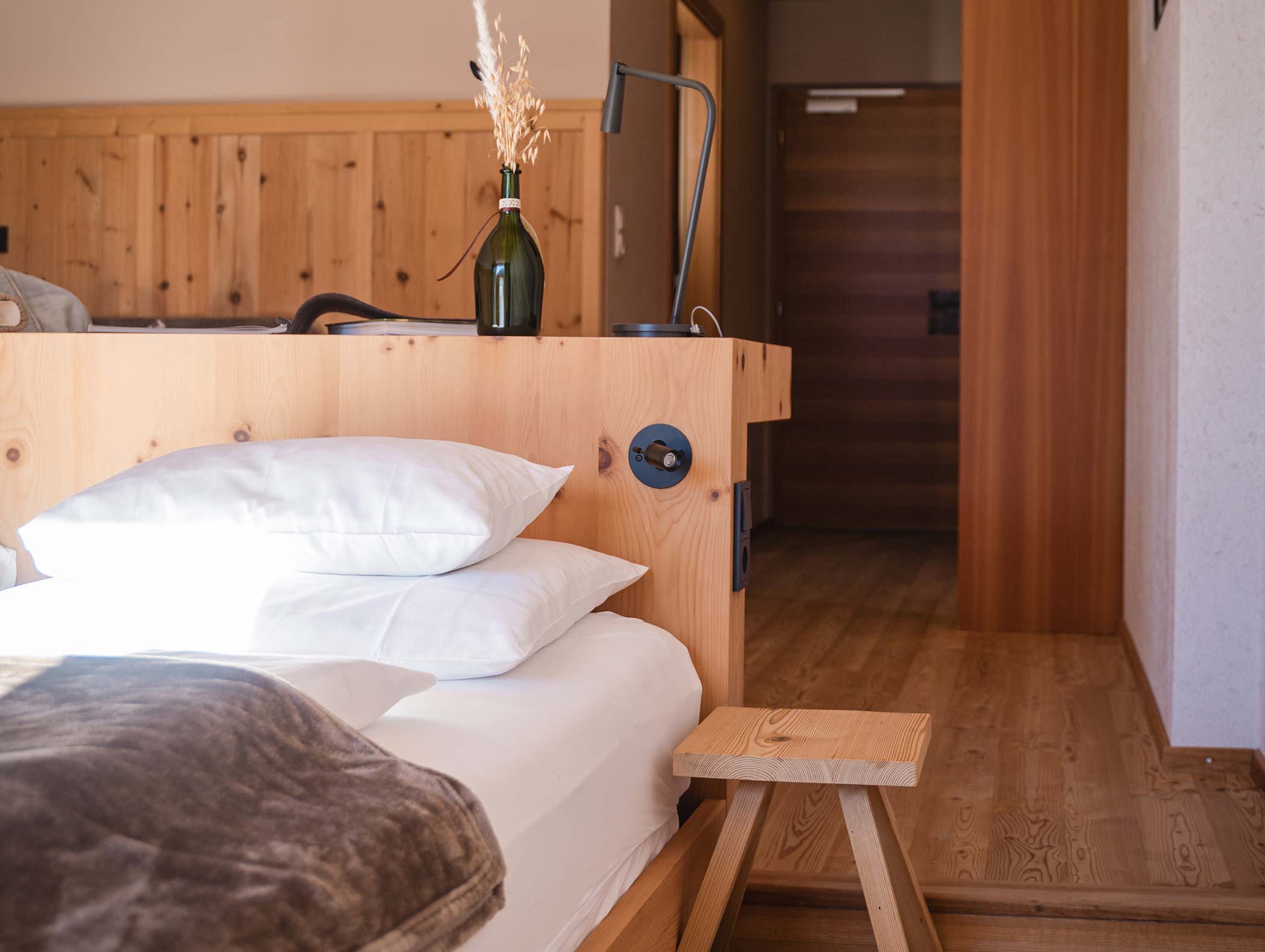 Accogliente camera da letto con mobili in legno, cuscini bianchi, una coperta e una bottiglia verde con fiori secchi. - Hotel Moosmair