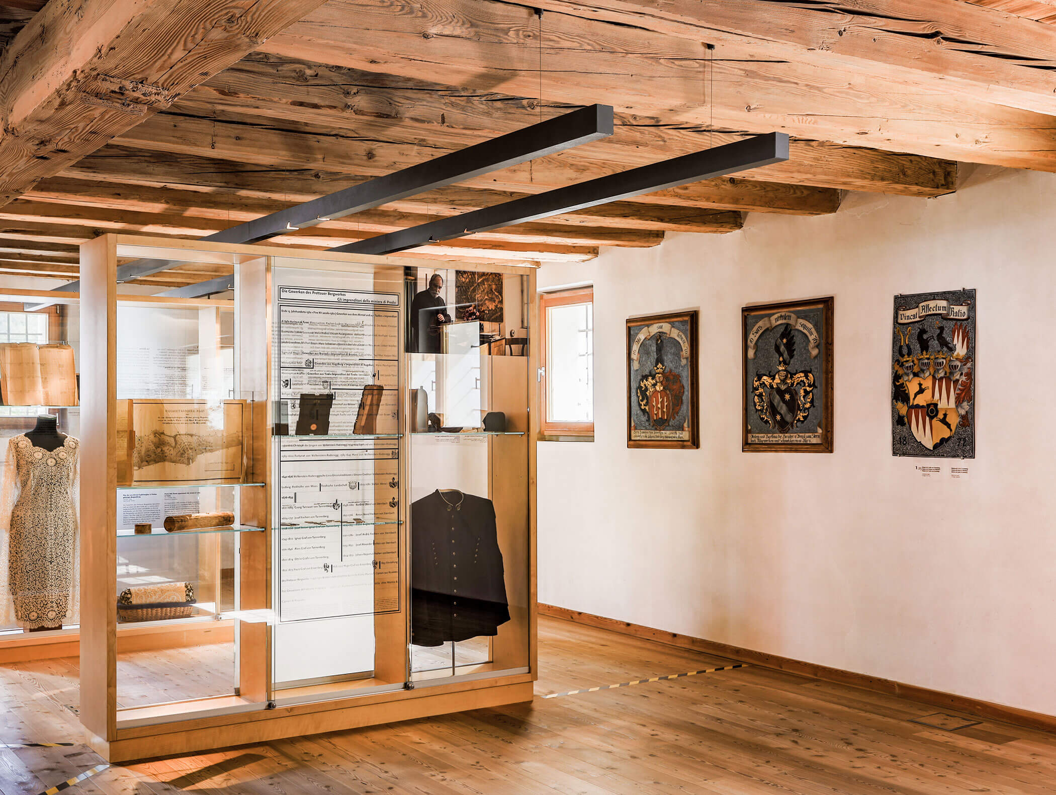 Esposizione museale con vetrina, abiti storici, libri e opere d'arte incorniciate su pareti di legno. - Hotel Moosmair