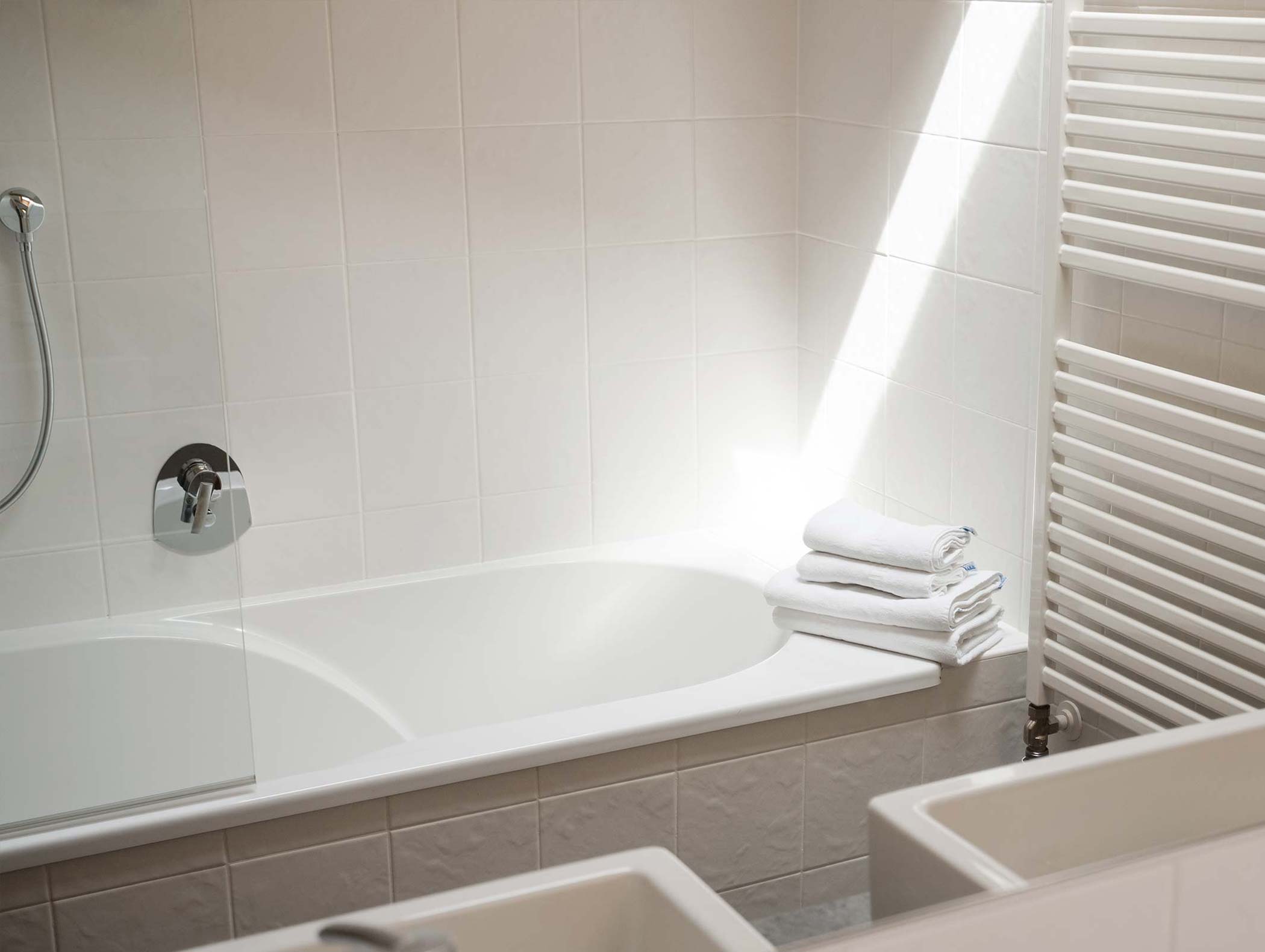Una vasca da bagno bianca con asciugamani piegati sul bordo, accanto a un radiatore in un bagno luminoso. - Hotel Moosmair