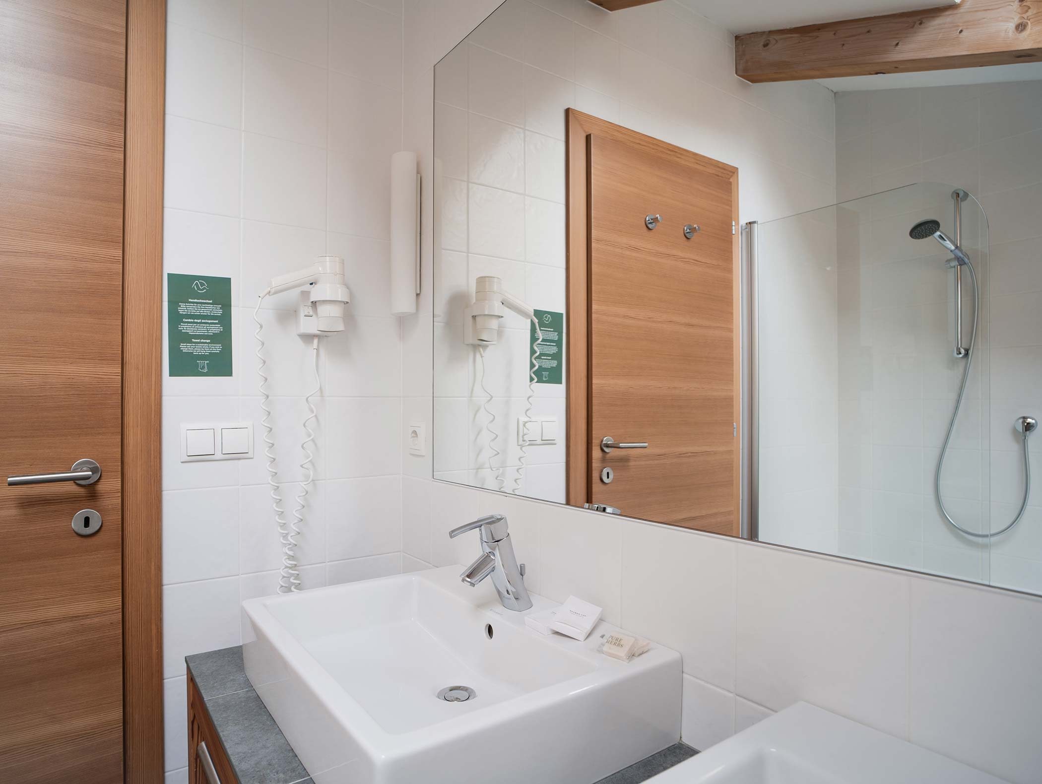 Bagno moderno con lavabo bianco, porta in legno, asciugacapelli a muro e cabina doccia in vetro. - Hotel Moosmair