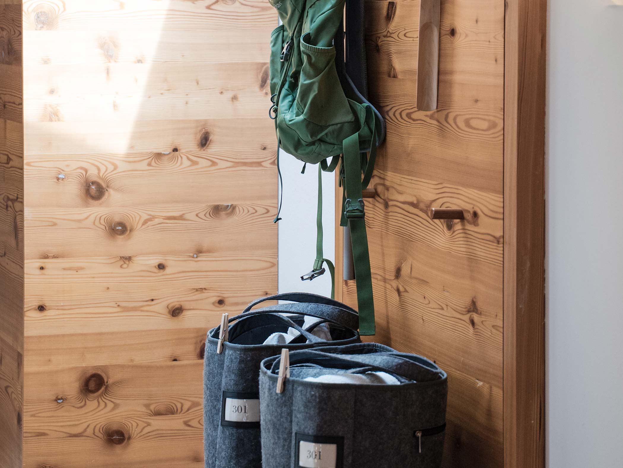 Uno zaino verde è appeso a una parete di legno sopra due cesti di feltro grigi pieni di coperte. - Hotel Moosmair