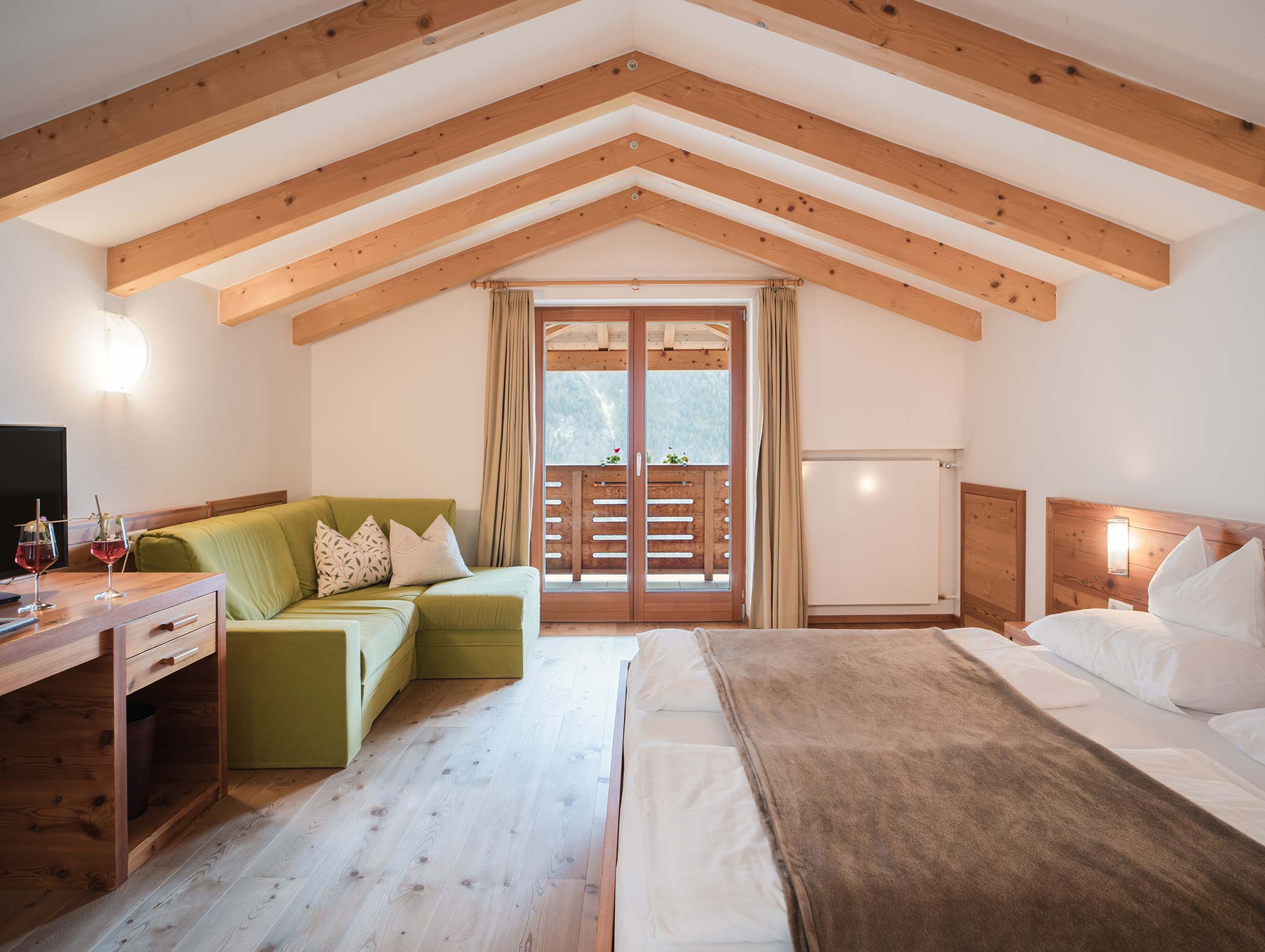 Accogliente camera da letto con travi in legno, divano verde, letto matrimoniale e porte del balcone che lasciano entrare la luce naturale. - Hotel Moosmair