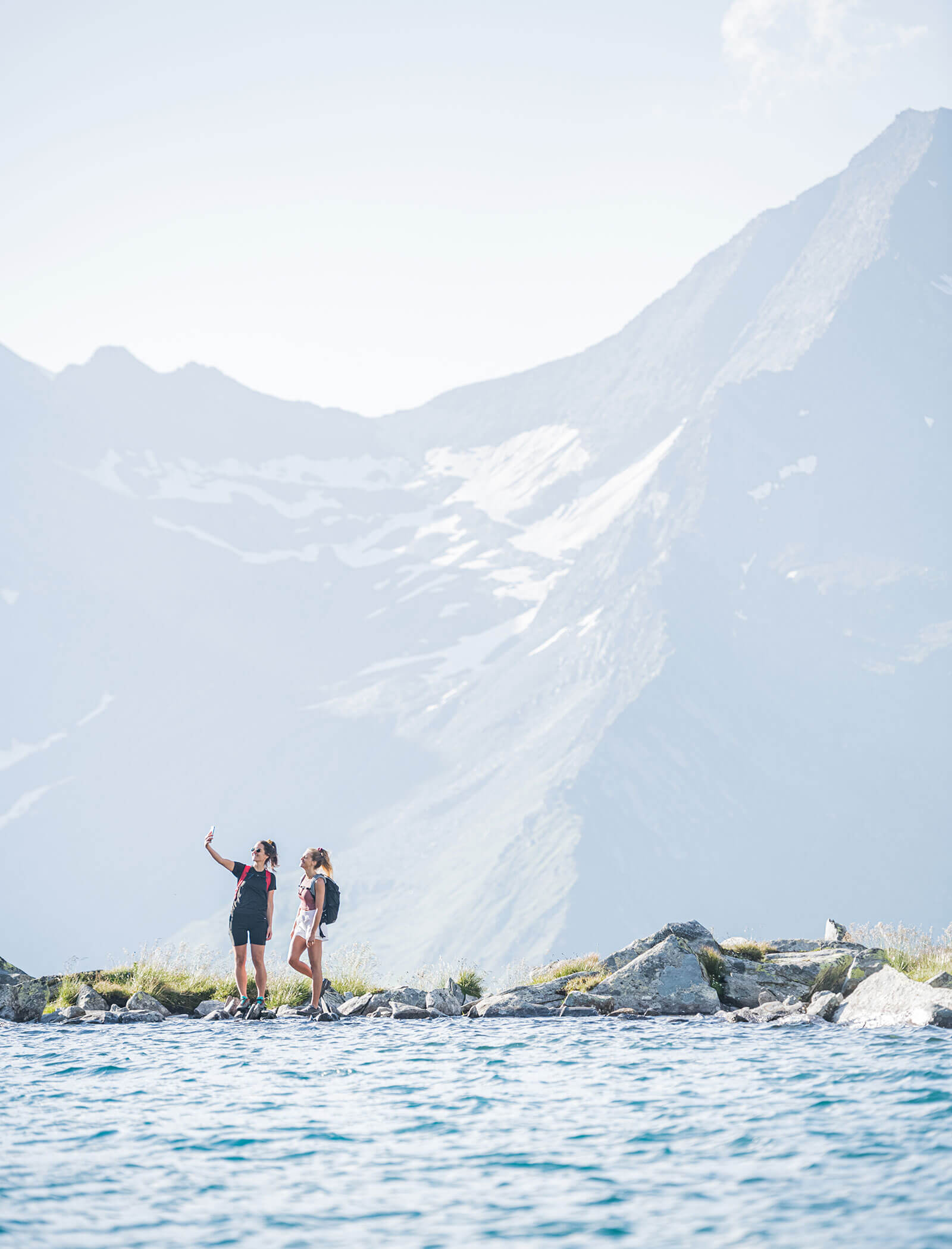 Two hikers taking a selfie beside a mountain lake with snowy peaks in the background. - Hotel Moosmair