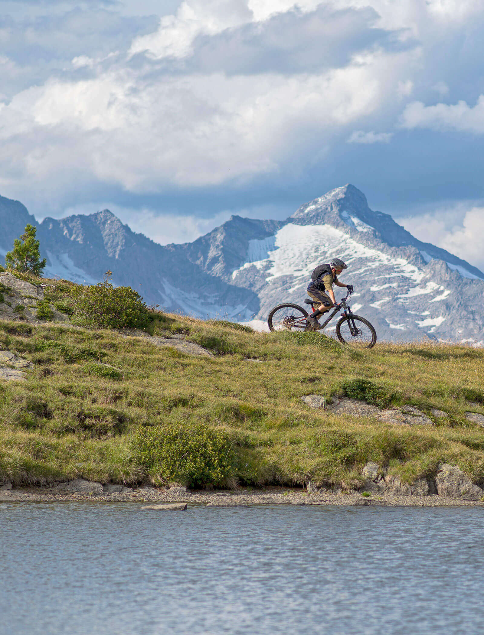 A cyclist rides a mountain bike on a grassy hill with snowy mountains and a lake in the background. - Hotel Moosmair
