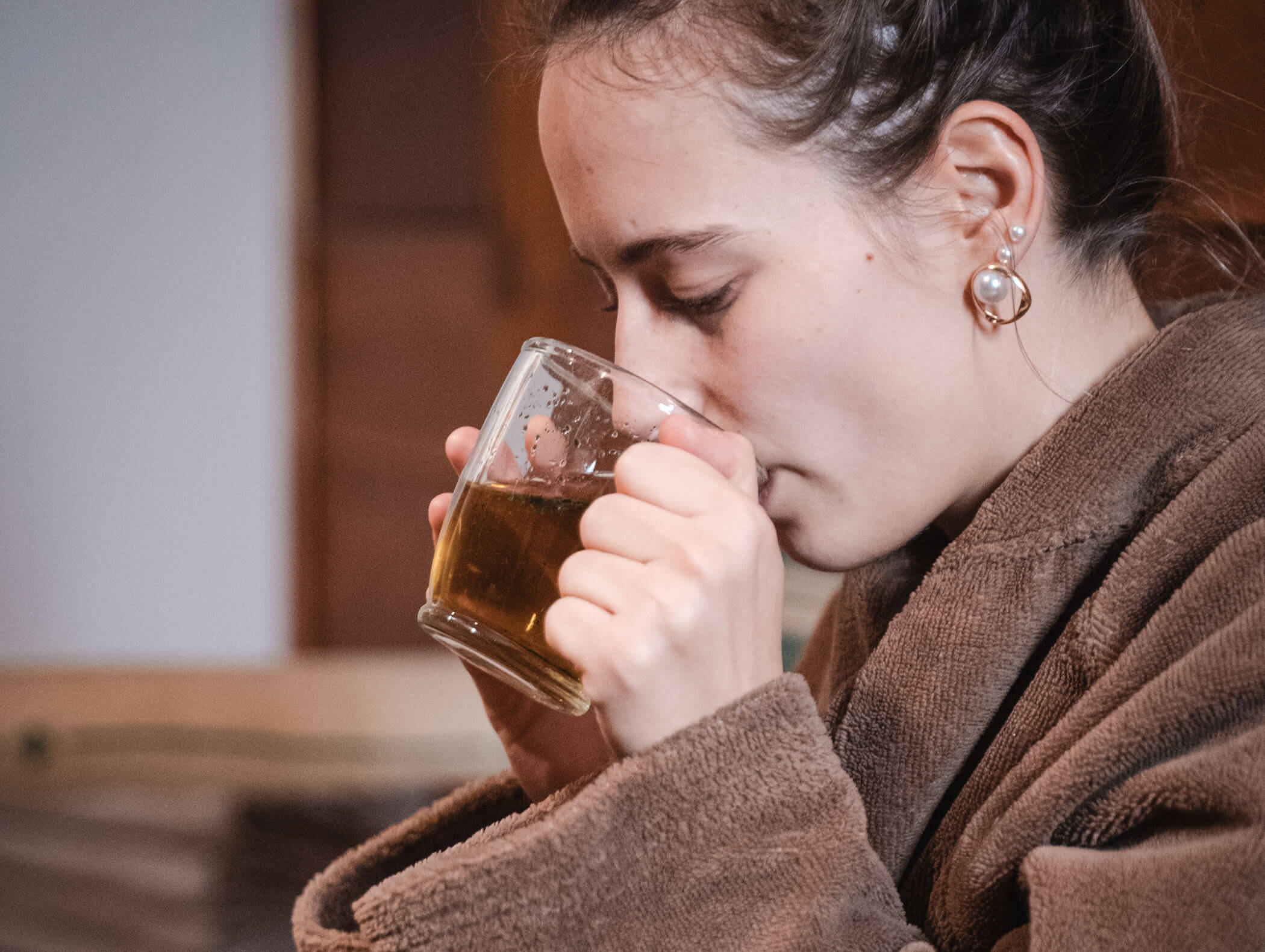 Donna in vestaglia marrone che beve da una tazza di tè in vetro, rivolta verso sinistra, in interni. - Hotel Moosmair