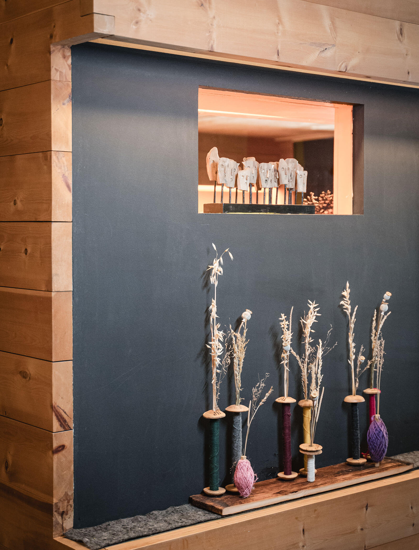 Wooden wall with a shelf holding dried plants in spools of thread, and ceramic faces in a window above. - Hotel Moosmair