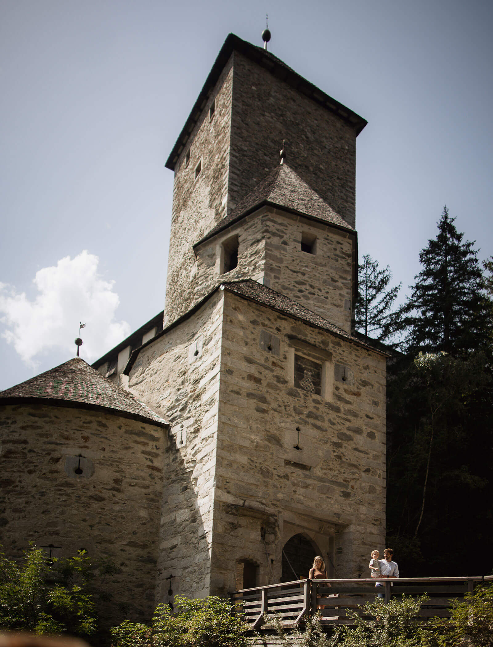 Two people stand on a wooden bridge in front of a tall stone tower surrounded by trees. - Hotel Moosmair