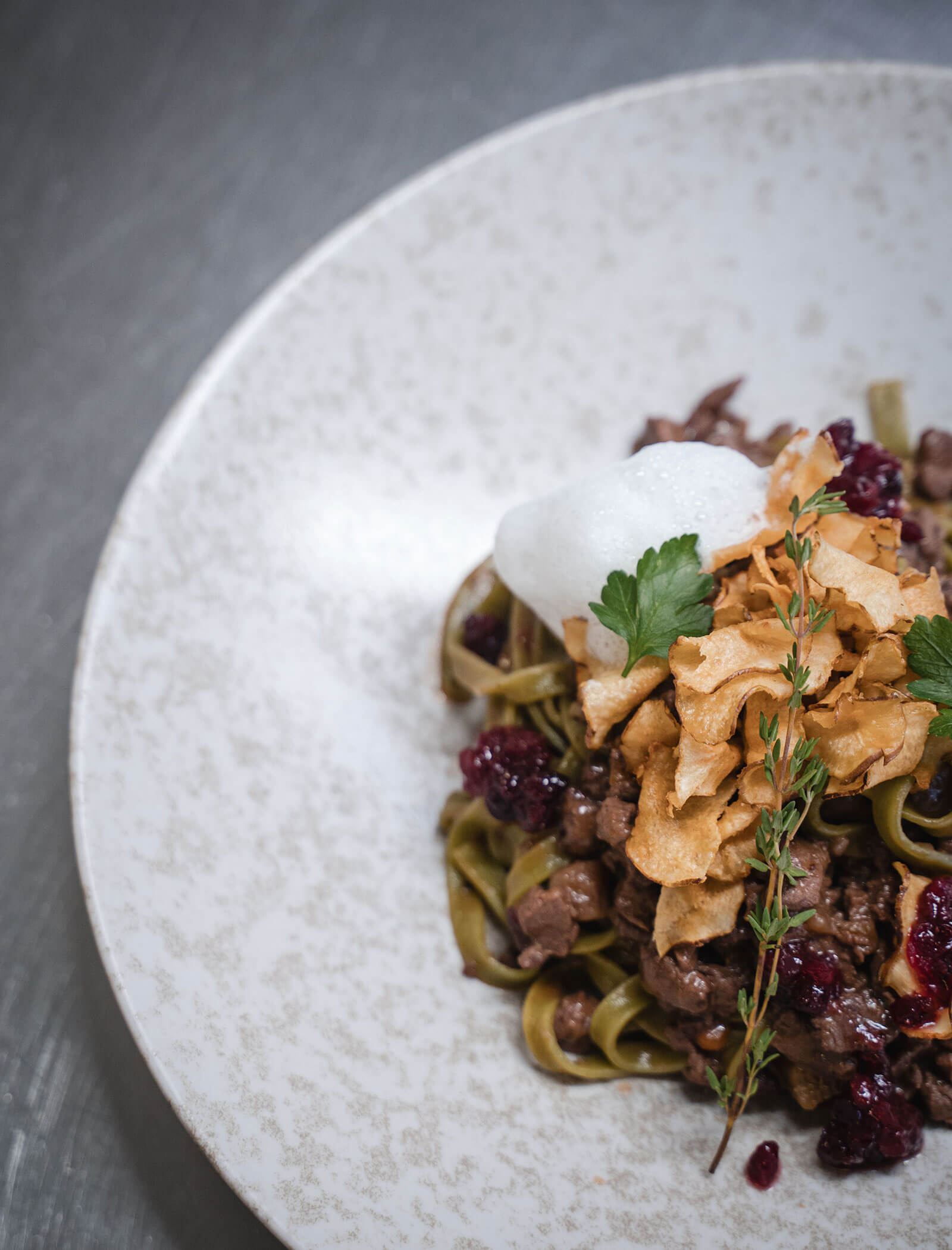 A plated dish of pasta topped with meat, crispy garnish, foam, herbs, and a berry sauce on a white plate. - Hotel Moosmair