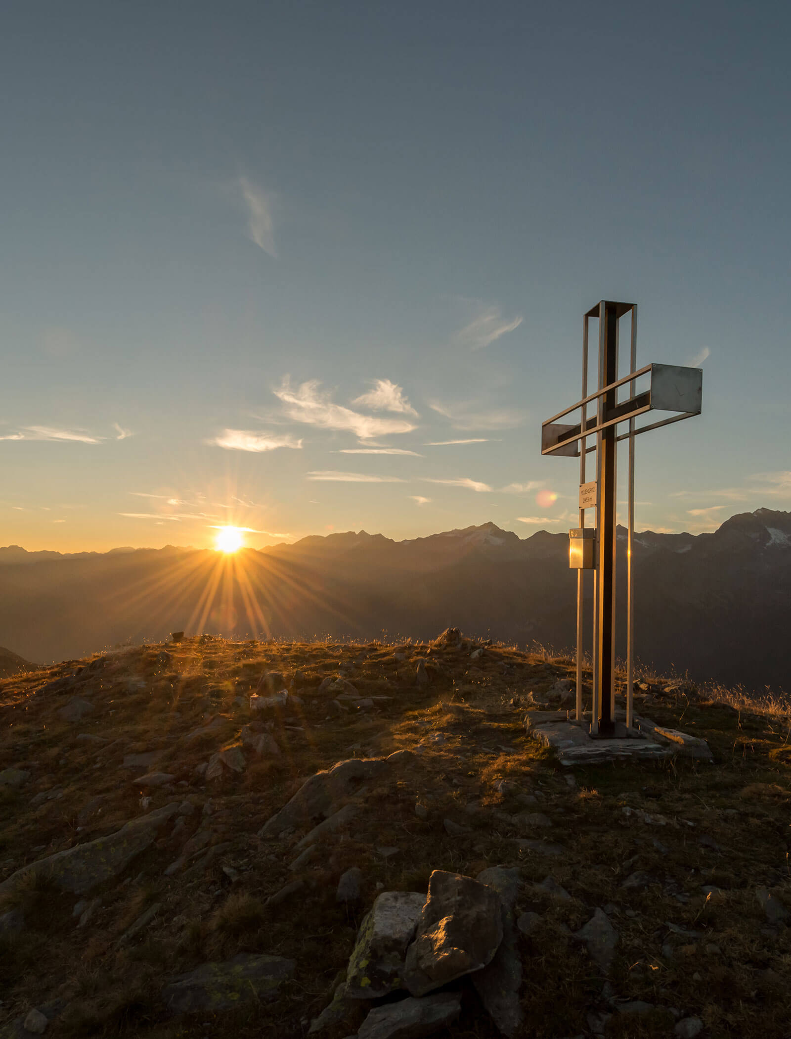 Croce di metallo sulla cima di una montagna con il sole che tramonta dietro le cime lontane in un cielo limpido. - Hotel Moosmair