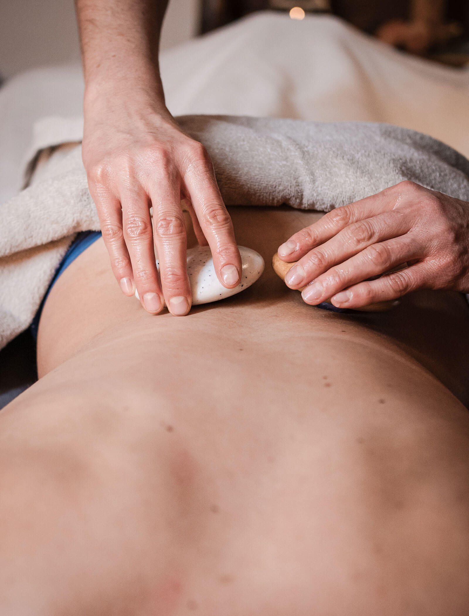 Mani che praticano un massaggio con pietre calde sulla schiena nuda di una persona, con un asciugamano che copre la parte inferiore del corpo. - Hotel Moosmair