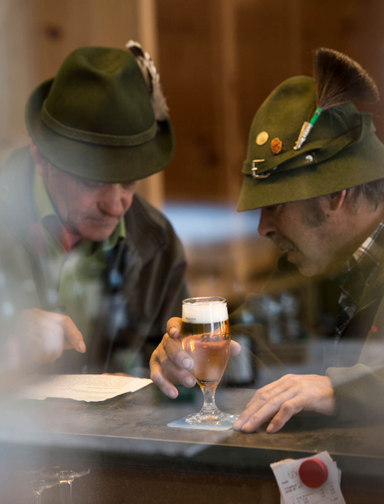 Due uomini con cappelli verdi e piume leggono insieme un giornale in un bar con un bicchiere di birra davanti a loro. - Hotel Moosmair