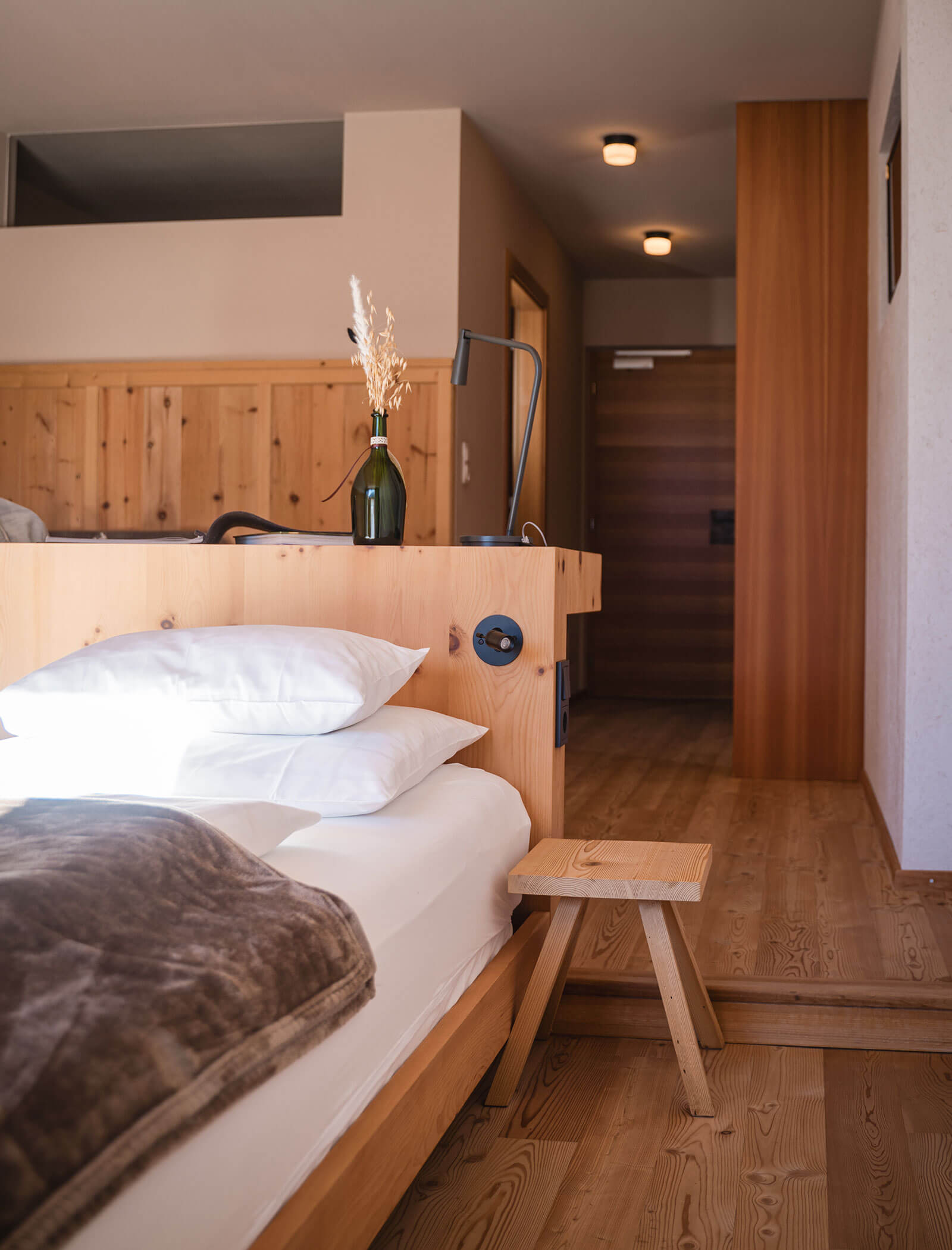 A cozy wooden hotel room with a neatly made bed, a small stool, and a vase with dried flowers. - Hotel Moosmair