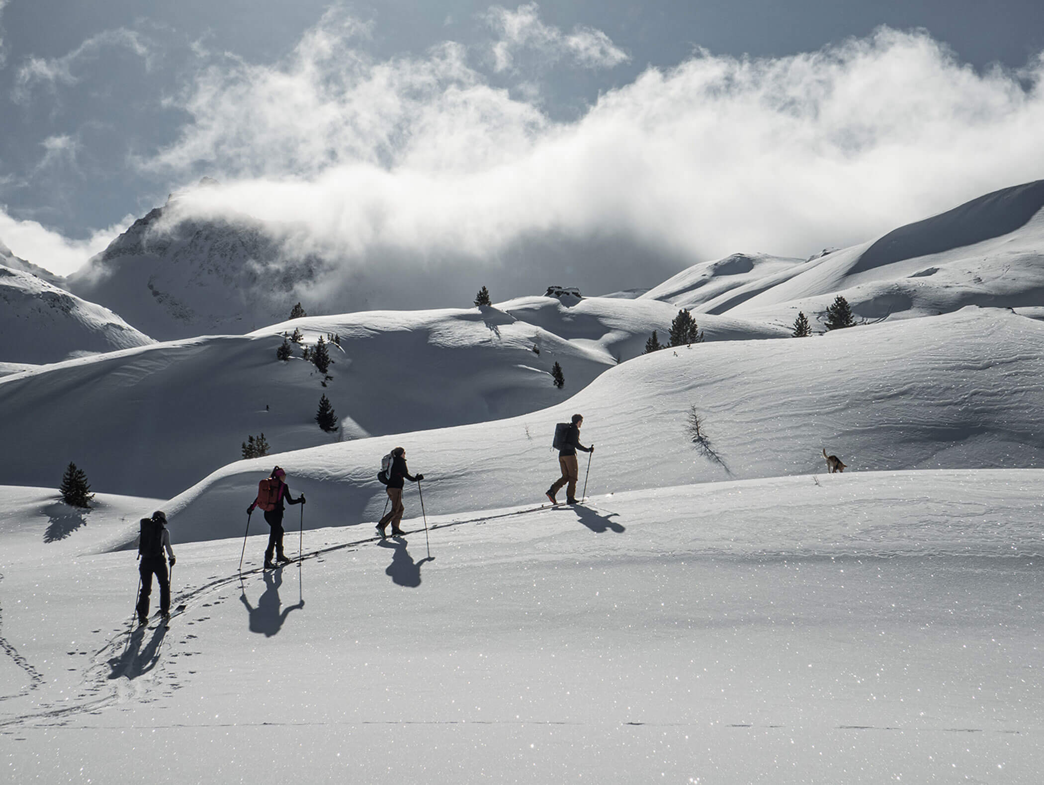 Quattro persone camminano con i bastoni su pendii innevati con montagne e nuvole sullo sfondo. - Hotel Moosmair