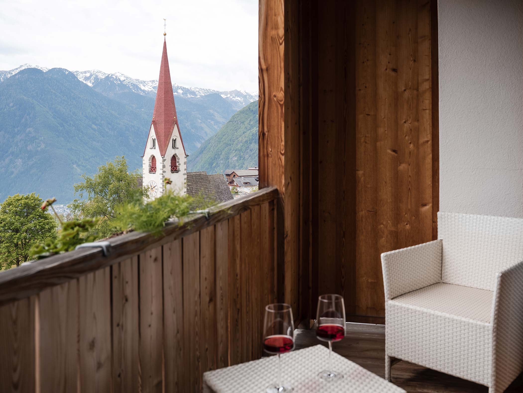 Due bicchieri di vino rosso su un balcone con vista sulle montagne e un campanile sullo sfondo. - Hotel Moosmair