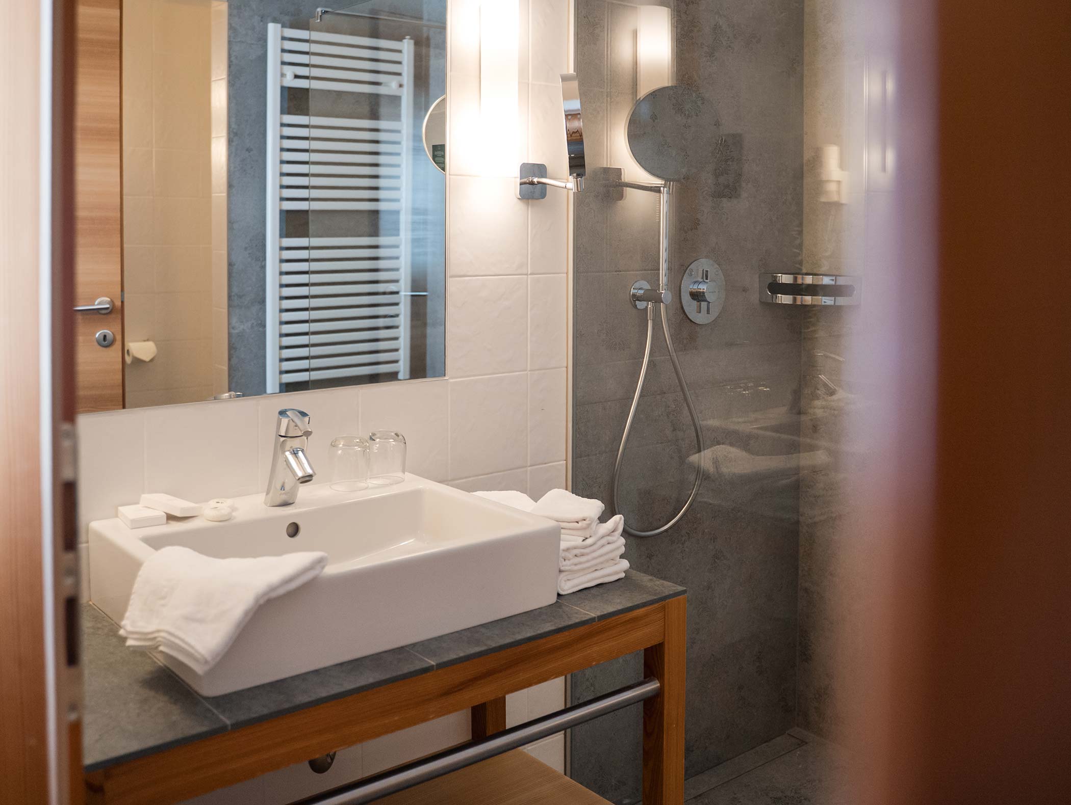 Bagno moderno con lavabo bianco, specchio, asciugamani e doccia in vetro con soffione a parete. - Hotel Moosmair