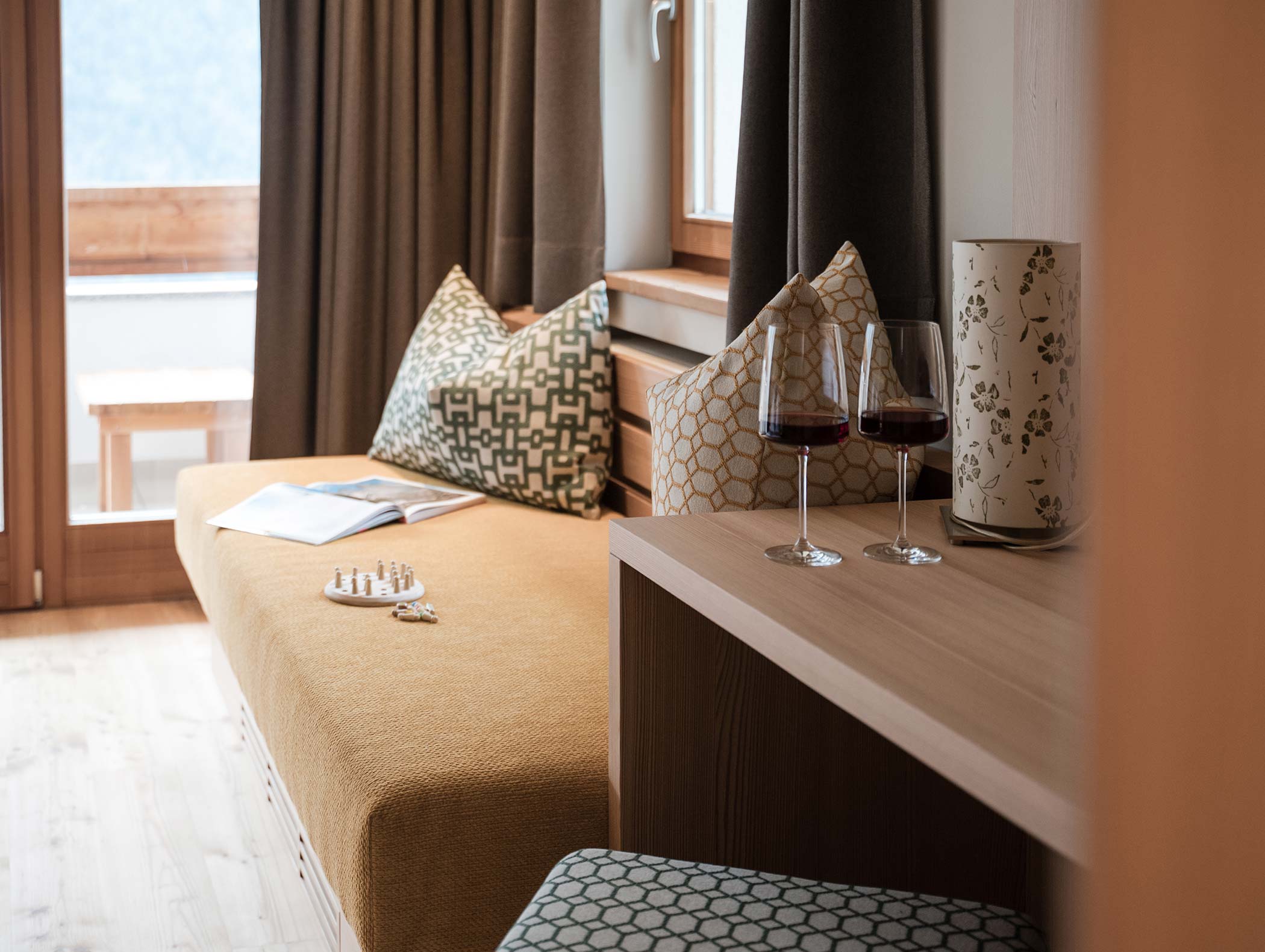 Un angolo accogliente con un lettino, cuscini, scacchi, un libro aperto e due bicchieri di vino rosso su un tavolo di legno. - Hotel Moosmair