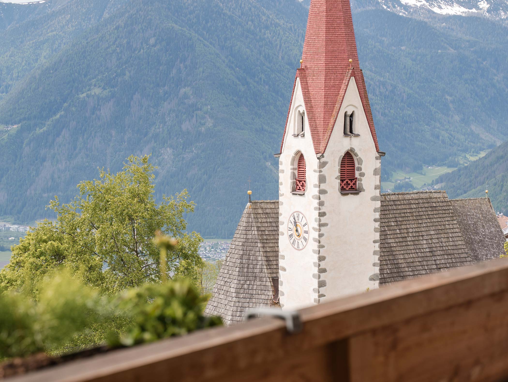 Church with a red steeple and clock, set against green mountains and trees in the background. - Hotel Moosmair