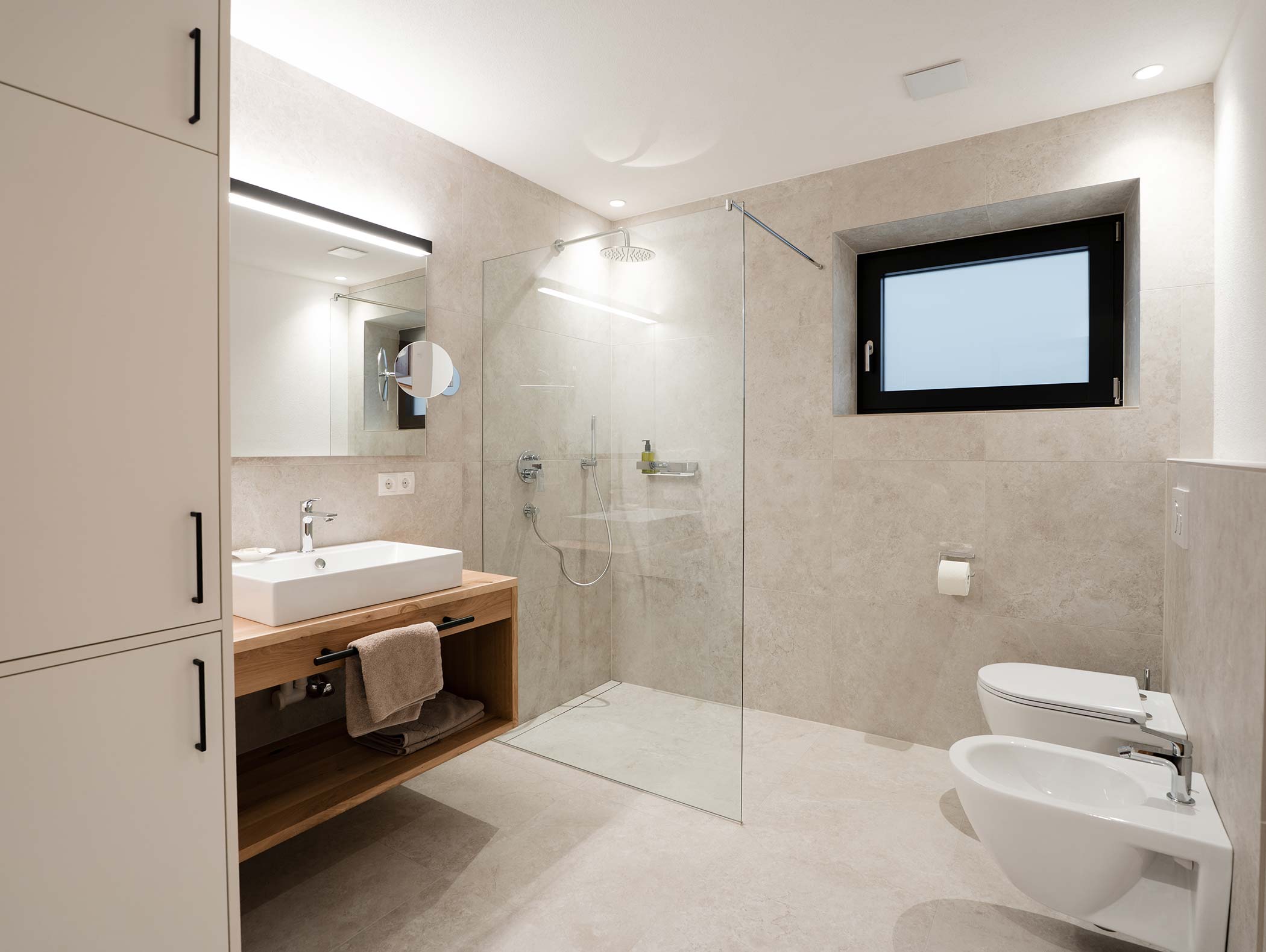 Bagno moderno con doccia in vetro, lavandino, bidet, WC, grande specchio e finestra sulla parete destra. - Hotel Moosmair