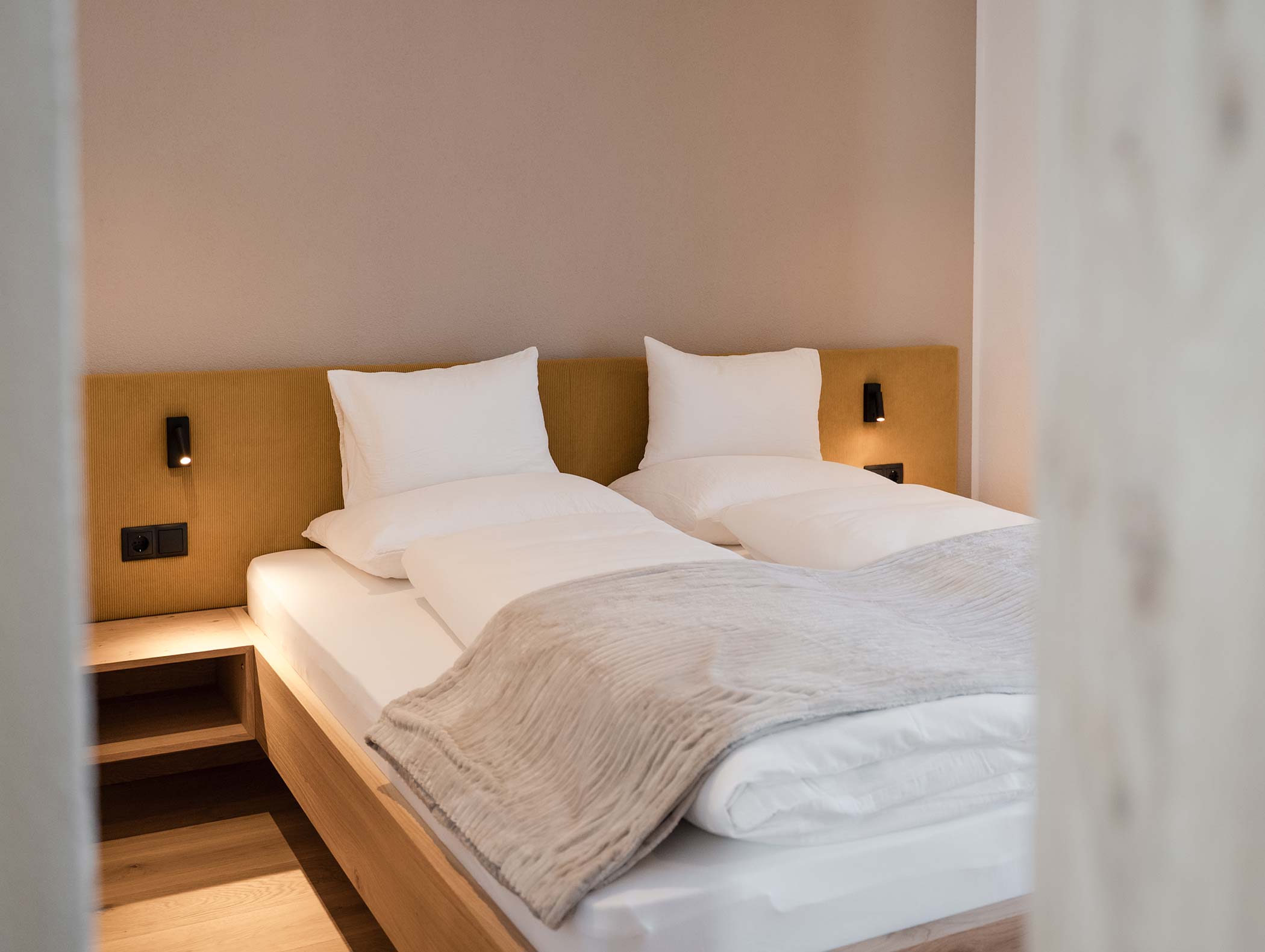Camera da letto minimalista con due letti gemelli, biancheria da letto bianca, testata del letto abbronzata e luci soffuse. - Hotel Moosmair