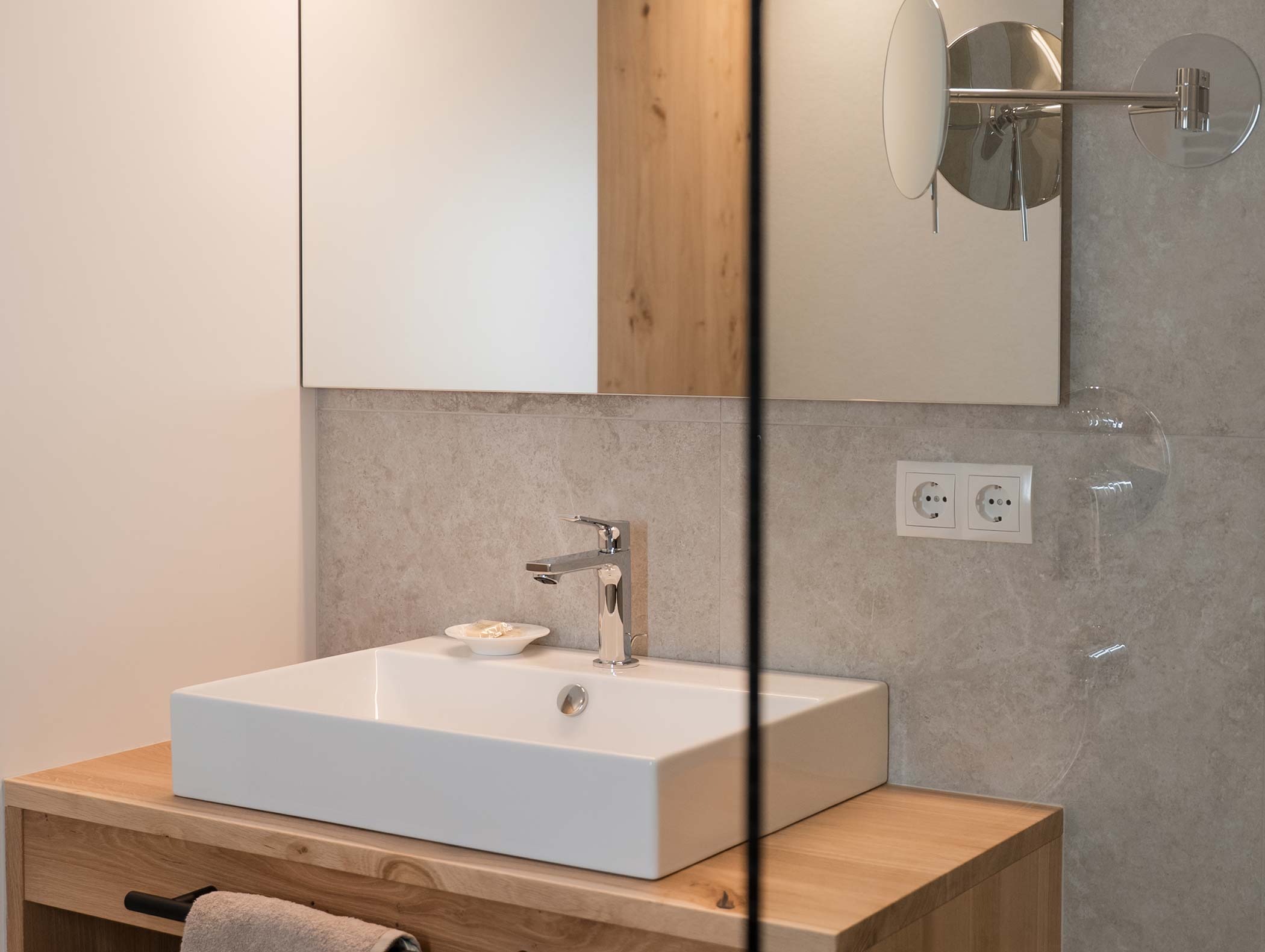 Bagno moderno con lavabo rettangolare, mobiletto in legno, specchio e prese a muro sopra un portasciugamani. - Hotel Moosmair