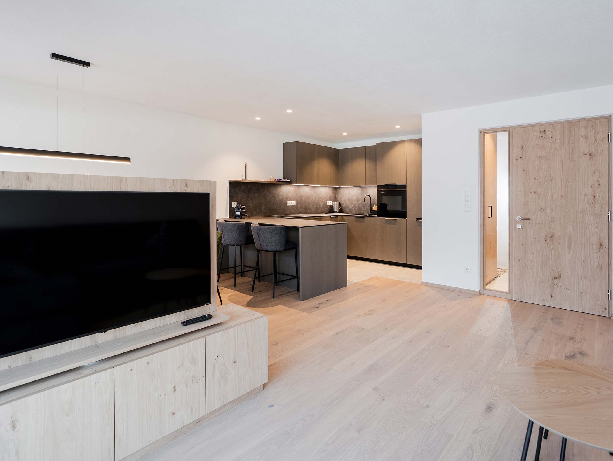 Moderno soggiorno e cucina a pianta aperta con pavimenti e armadi in legno, TV e isola con sgabelli. - Hotel Moosmair