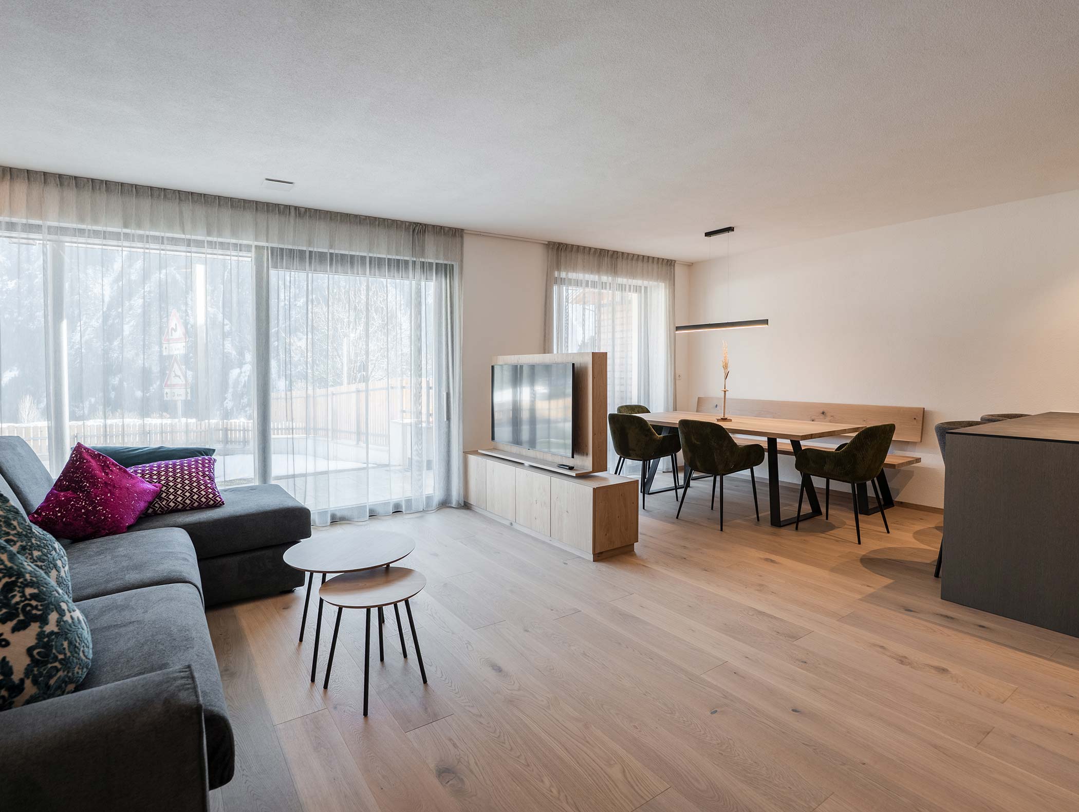 Moderna zona giorno e pranzo con grandi finestre, divano grigio, TV, pavimenti in legno e sedie verdi. - Hotel Moosmair