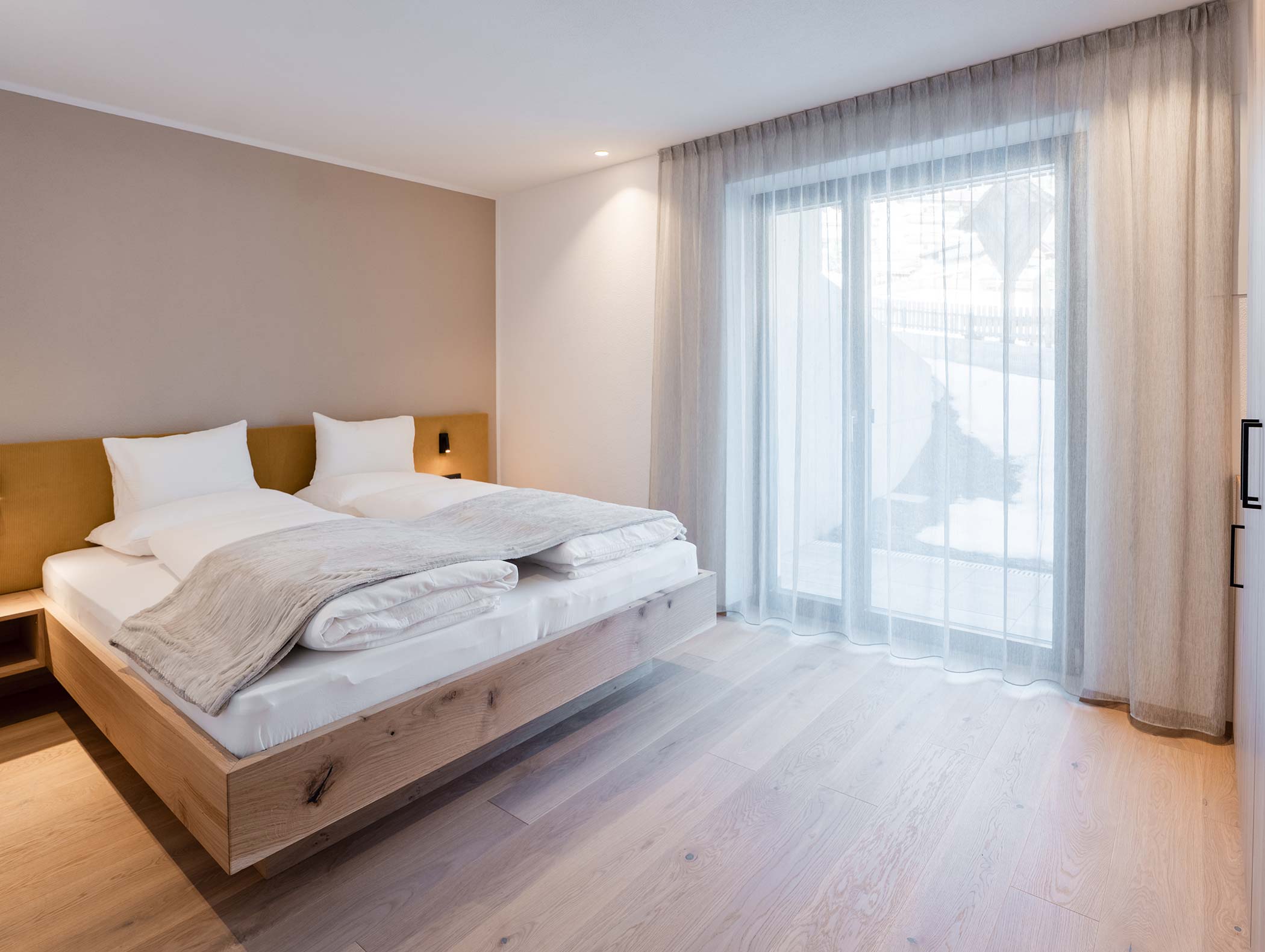 Camera da letto minimalista con letto grande, pavimento in legno chiaro e tende velate che coprono una finestra verso l'esterno. - Hotel Moosmair