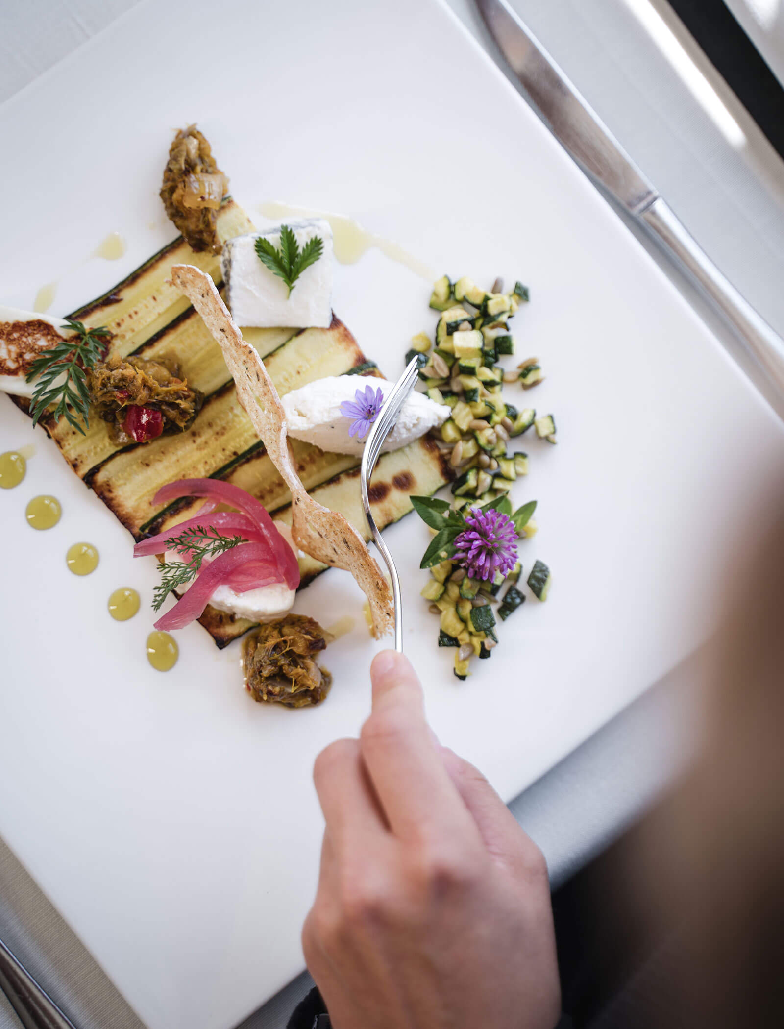 Eine Hand hält eine Gabel über einen Gourmet-Teller mit gegrilltem Gemüse, Käse, Kräutern und essbaren Blumen. - Hotel Moosmair