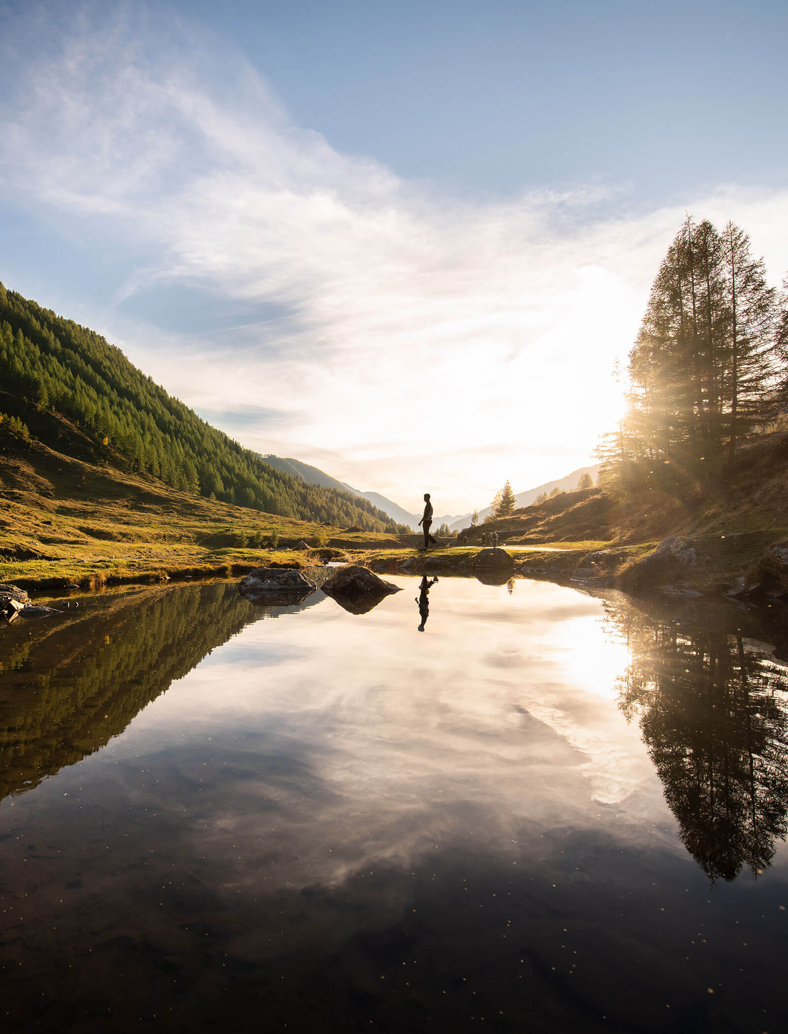 Person, die an einem ruhigen See steht, in dem sich Berge und Bäume bei Sonnenuntergang spiegeln. - Hotel Moosmair