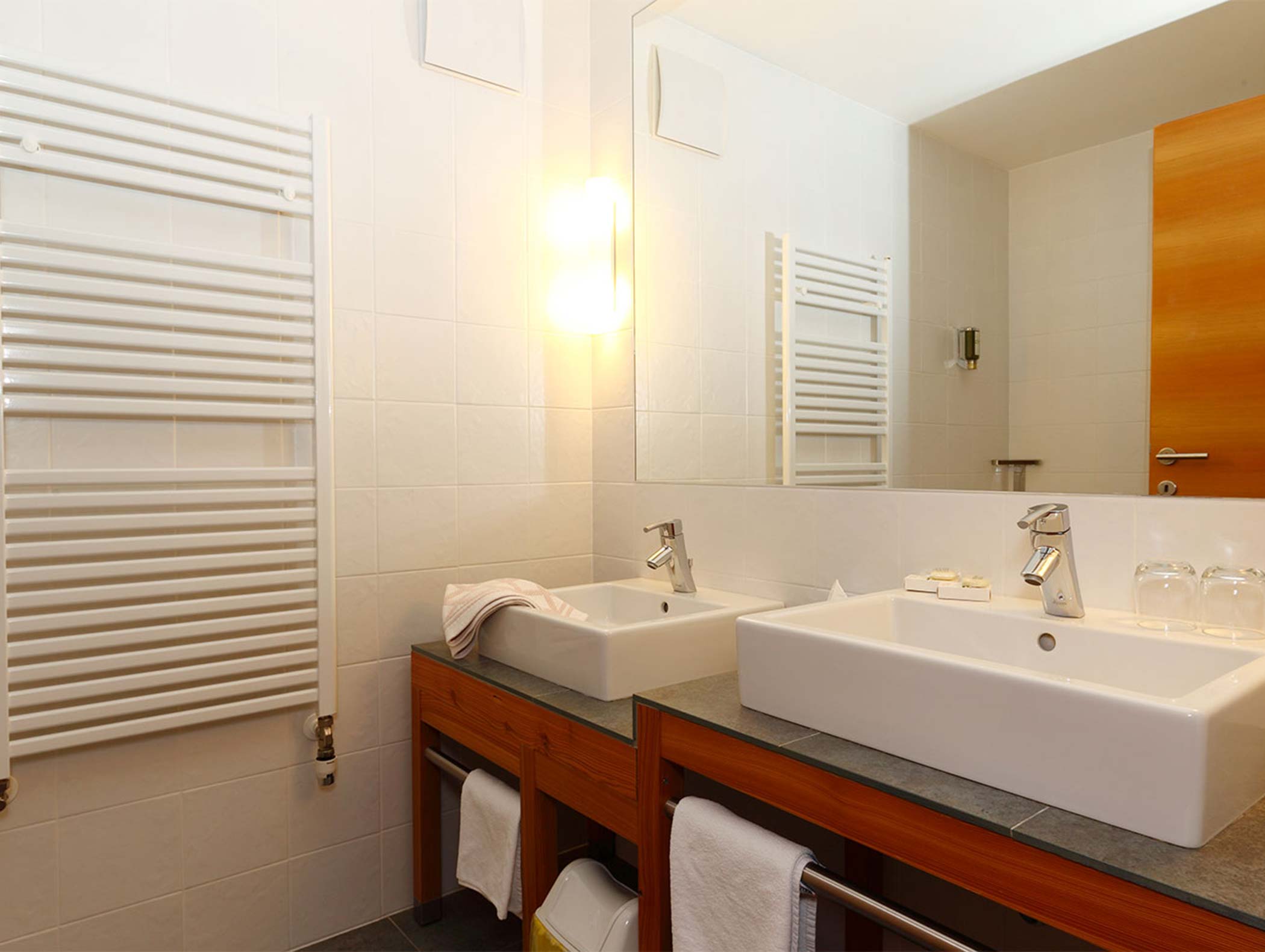 Bagno moderno con due lavandini bianchi, un mobile in legno, un grande specchio, un portasciugamani e una luminosa applique. - Hotel Moosmair