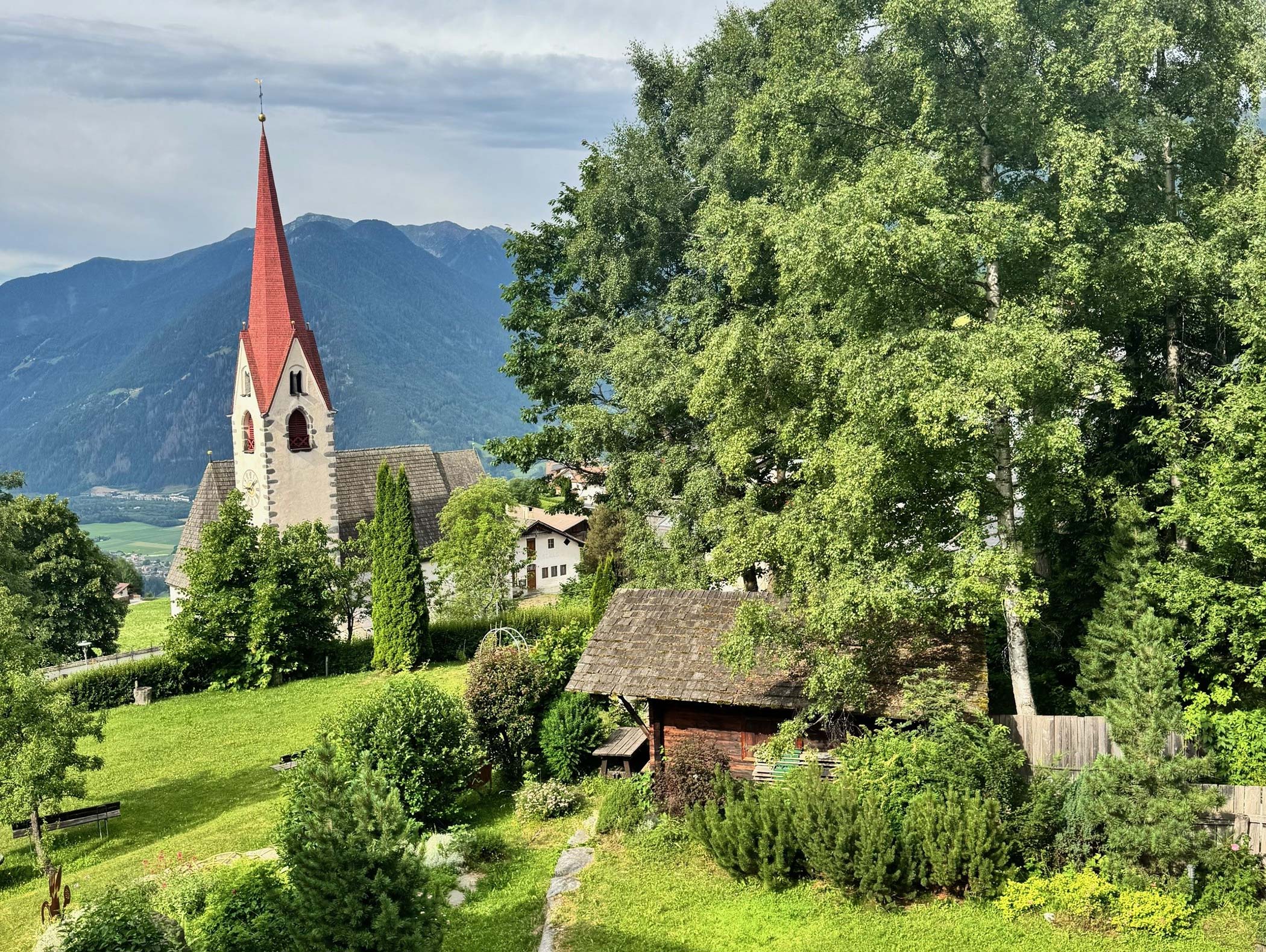 Una chiesa con un campanile rosso si erge tra gli alberi e le case, con alle spalle le montagne sotto un cielo parzialmente nuvoloso. - Hotel Moosmair