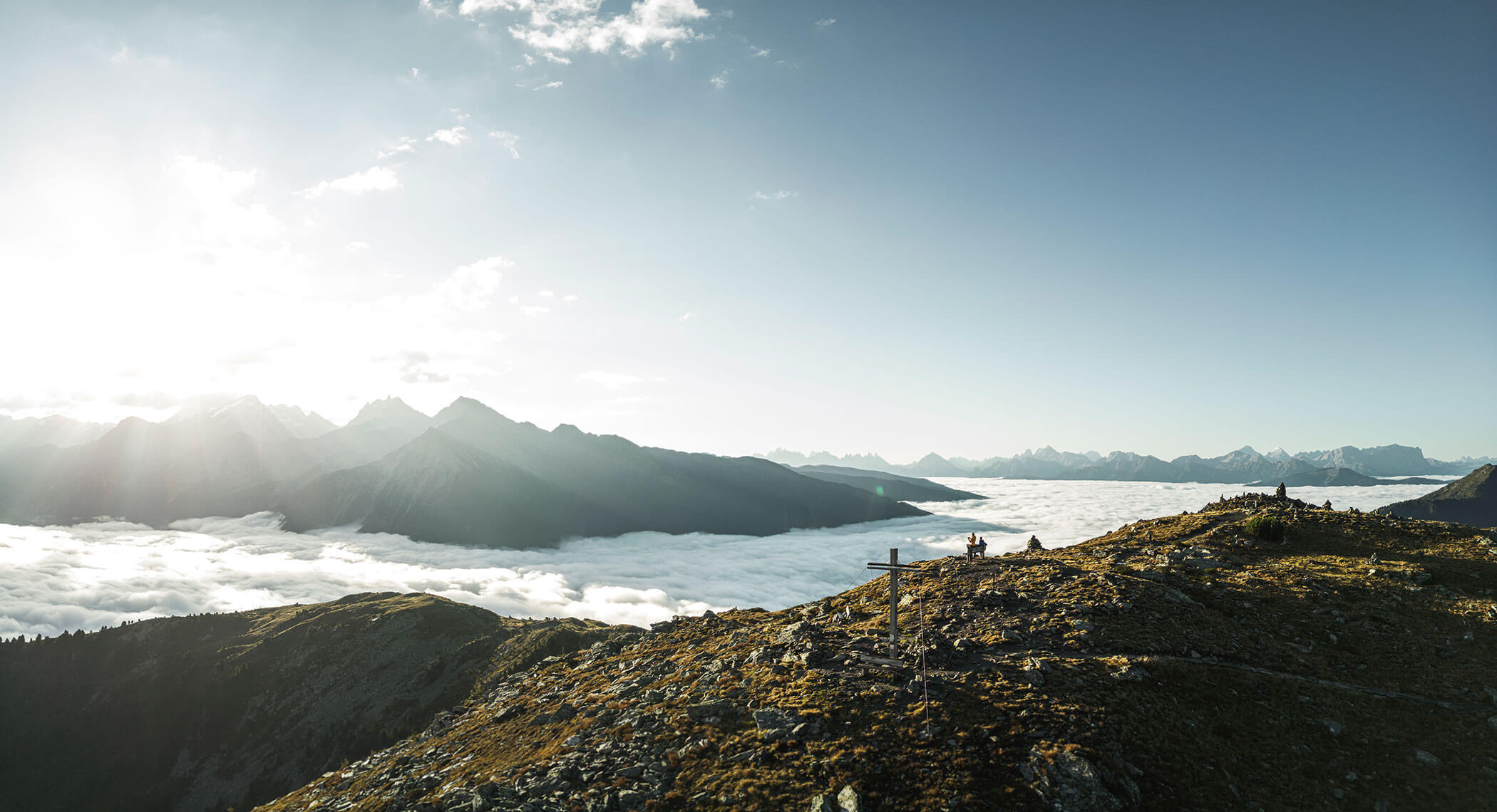 Mountain ridge above a sea of clouds with distant peaks and hikers near a wooden cross in sunlight. - Hotel Moosmair
