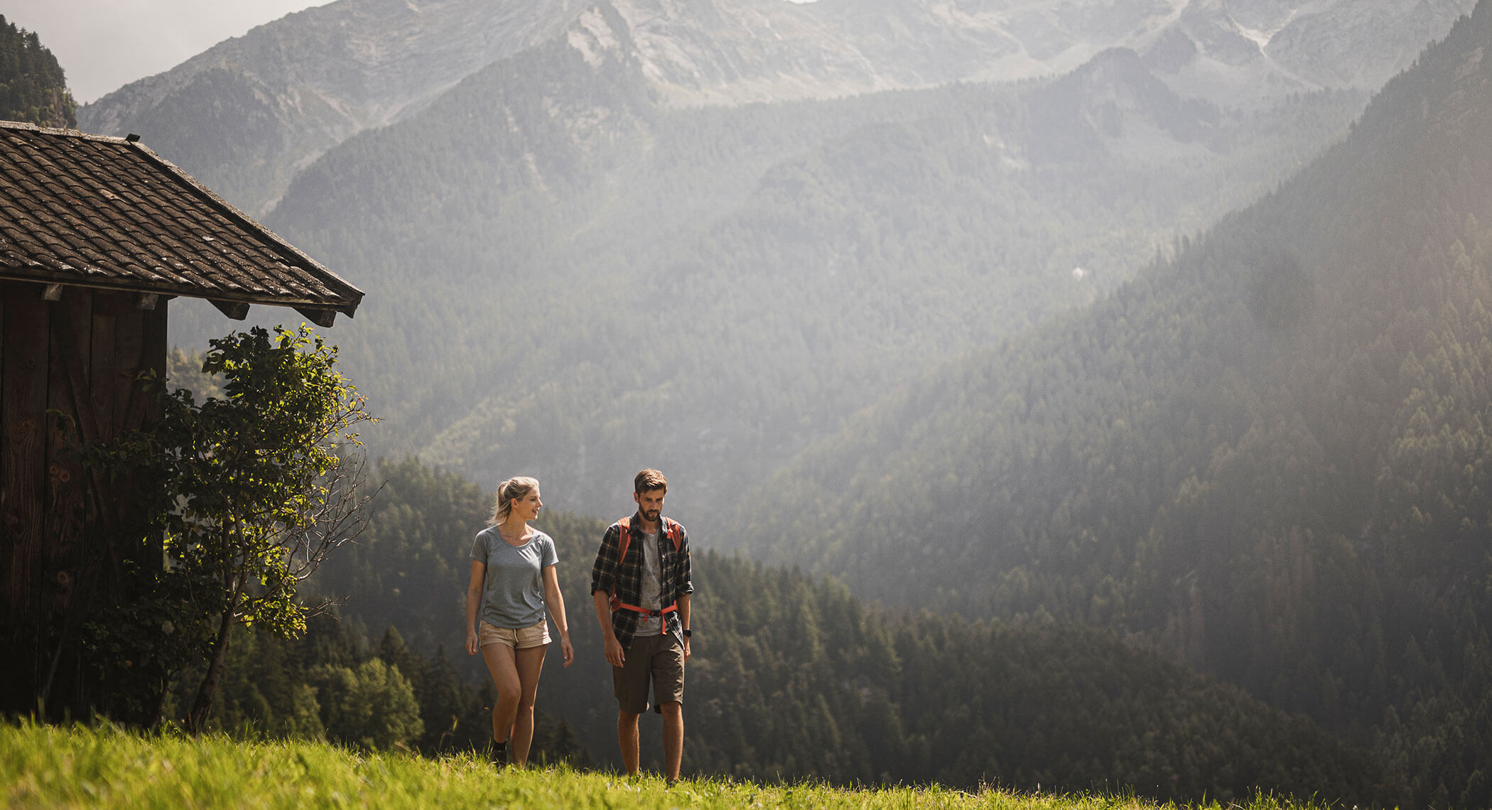 Ein Paar wandert auf einem grasbewachsenen Hügel neben einer Hütte mit bewaldeten Bergen im Hintergrund. - Hotel Moosmair