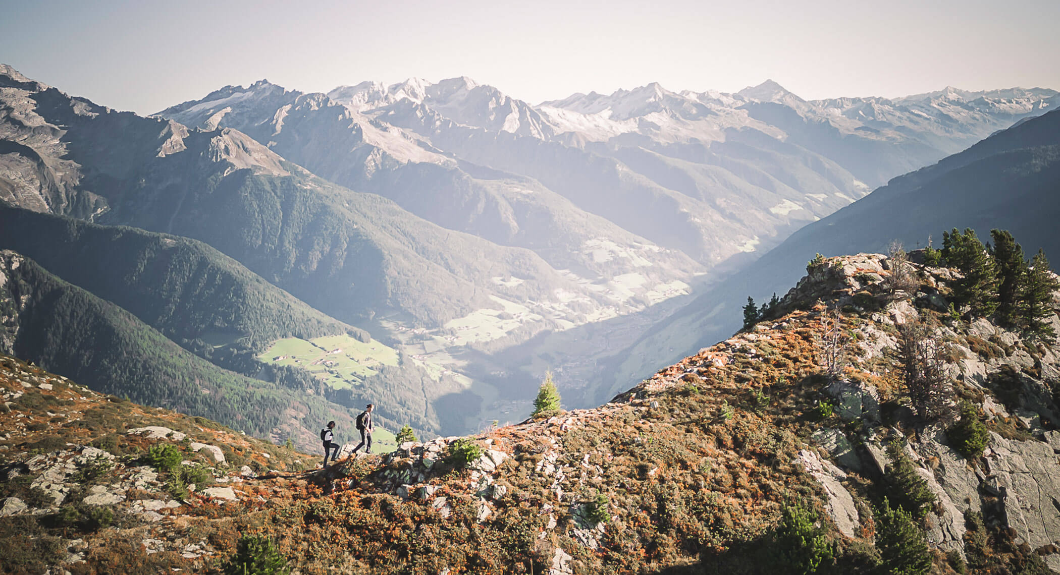 Two hikers walk along a mountain ridge with tall, snow-capped peaks in the background under clear skies. - Hotel Moosmair
